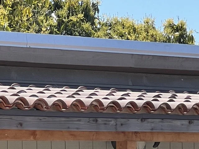 Close-up of a house roof with terracotta tiles and wooden trim, with green trees and blue sky in the background