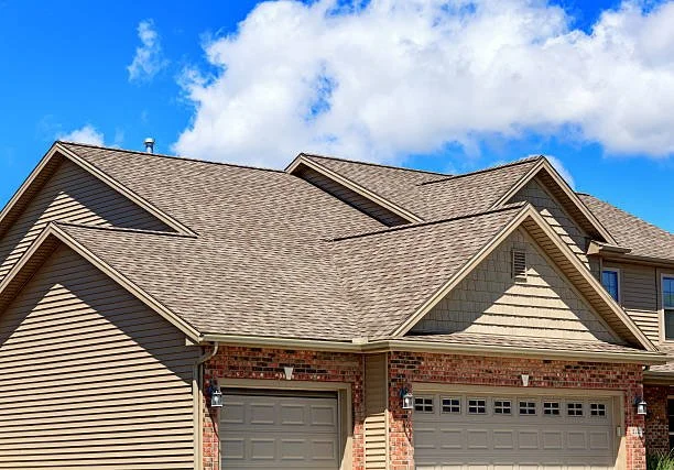 A residential house with a beige roof, siding, and brick accents, featuring a two-car garage and a partly cloudy blue sky.