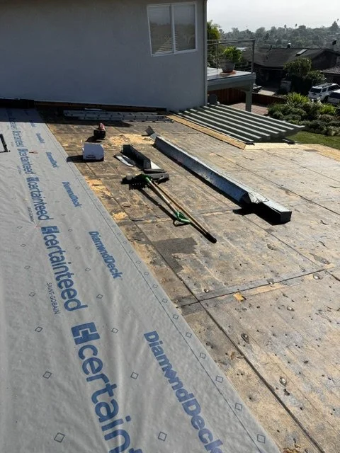 Roof under construction with tools, wooden planks, and insulation material, with a house and urban neighborhood in the background.
