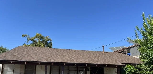 View of a house with a brown roof and a clear blue sky, with some trees and a small structure with solar panels visible in the background.