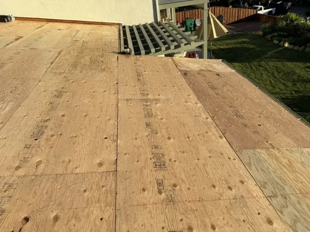 A roof under construction with plywood sheets nailed down, overlooking a backyard with a deck and green lawn.