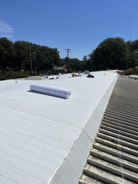 View of a white flat rooftop with roofing materials and tools, trees, and power lines in the background under a clear blue sky.