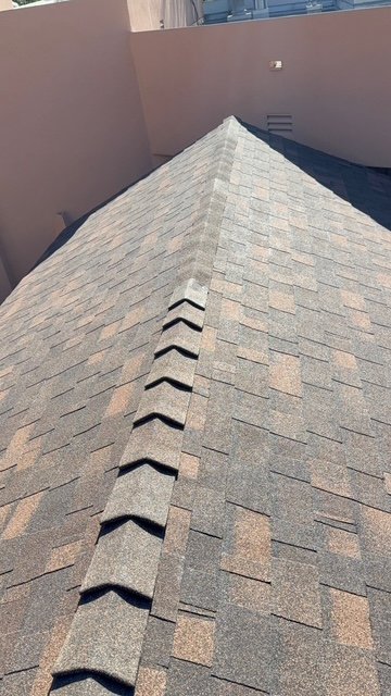 View of a house roof with gray asphalt shingles and ridge vent, surrounded by beige walls.