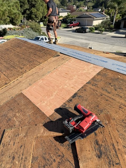 A worker standing on a roof under construction, with a nail gun and several sheets of plywood, as part of roofing work in progress.