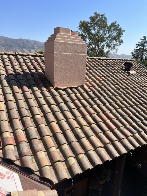View of a tiled roof with a large chimney and a smaller vent, with mountains and trees in the background under a clear blue sky.