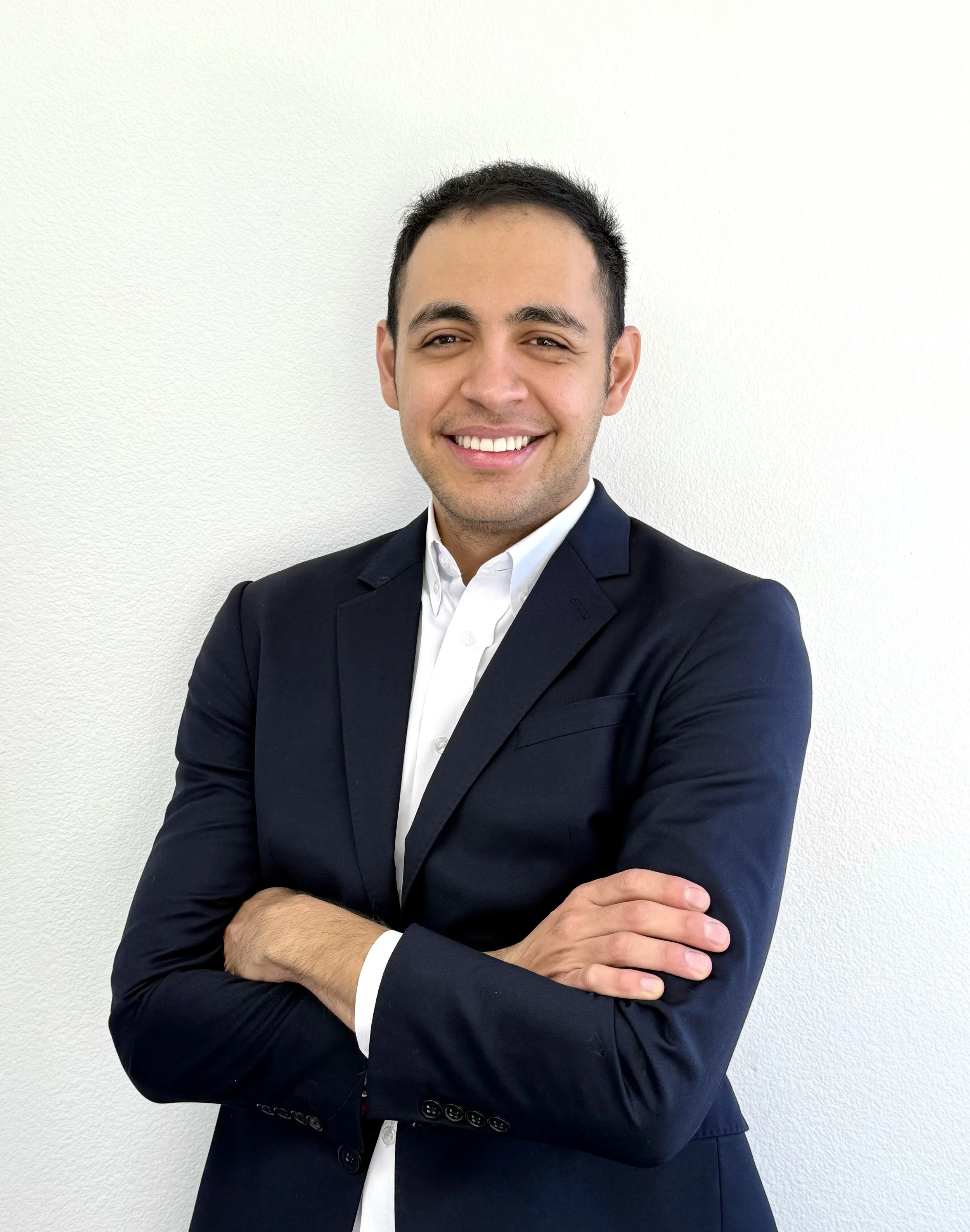 A man in a dark suit and white shirt smiling with crossed arms, standing against a plain light-colored wall.