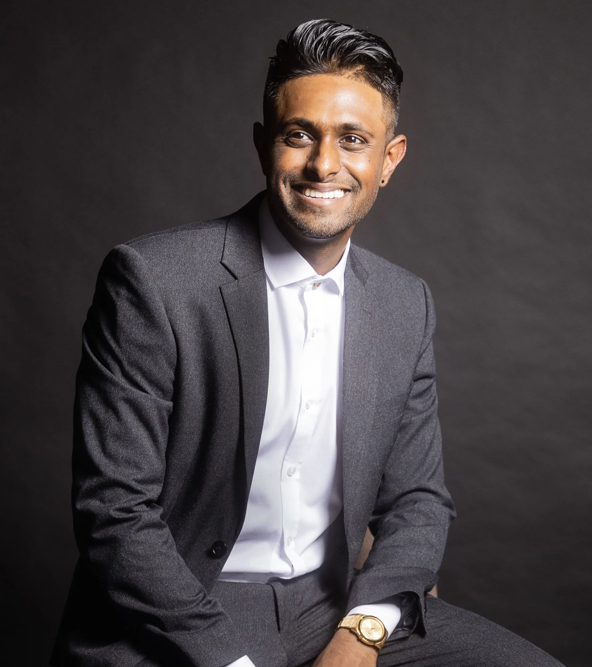 Portrait of a smiling man in a gray suit, white shirt, wearing a gold watch, posed against a dark background.
