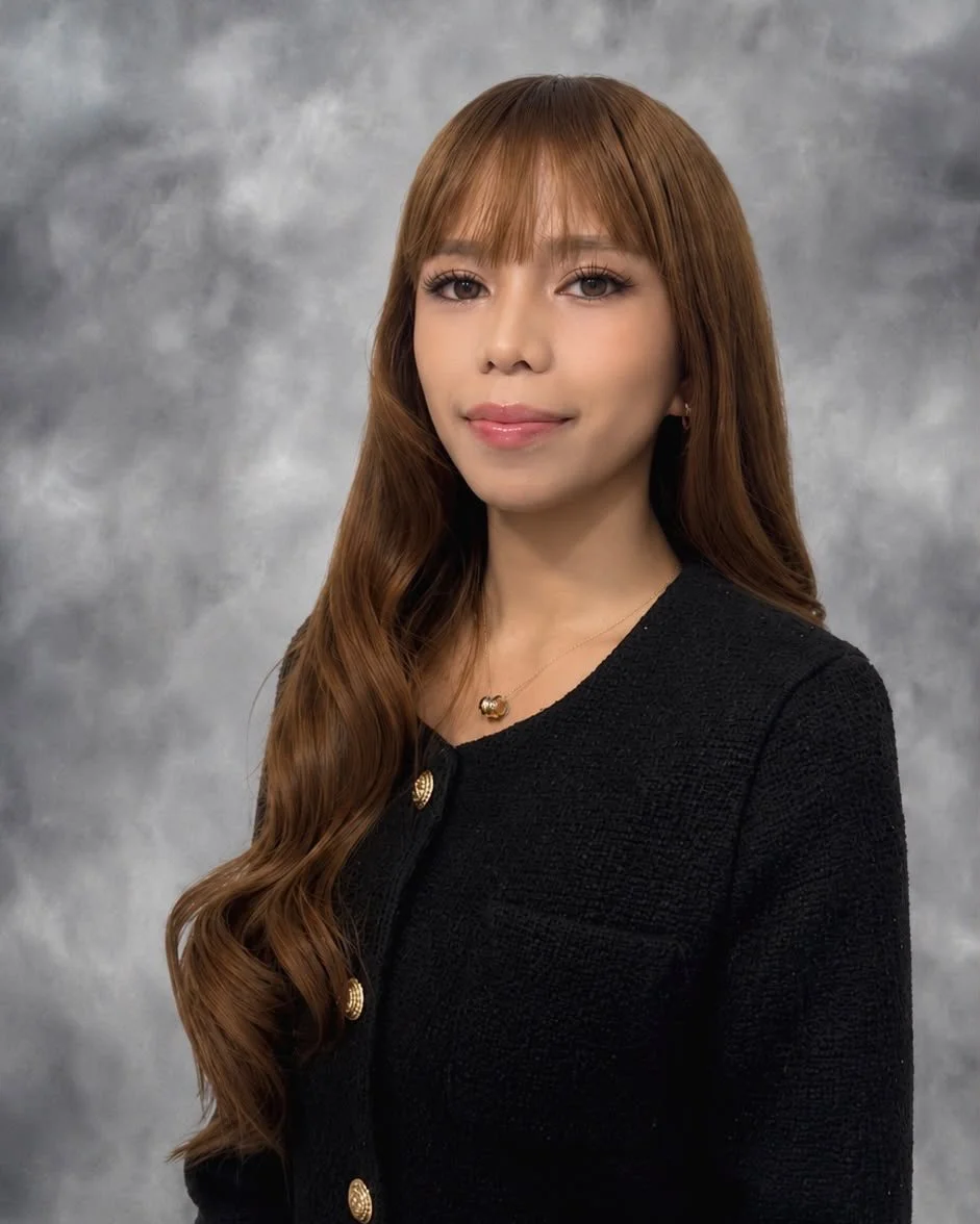 A young woman with long, wavy reddish-brown hair and light makeup, wearing a black textured blazer with gold buttons and a gold necklace, standing against a gray textured background.