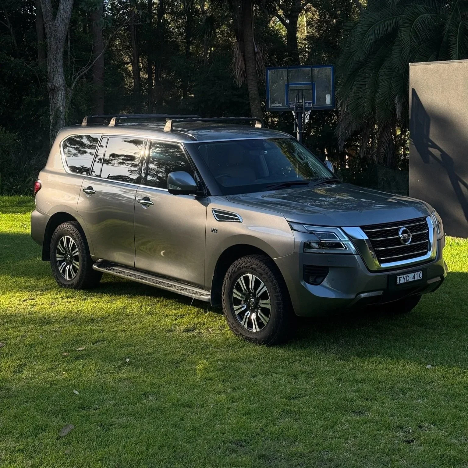A silver Nissan SUV parked on green grass with a basketball hoop and trees in the background.