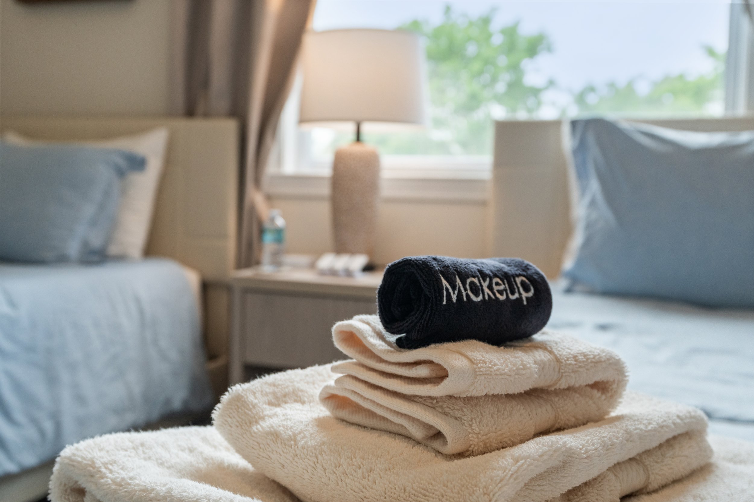 Stacked white and beige towels with a rolled black towel that has 'Makeup' written on it, on a bed.