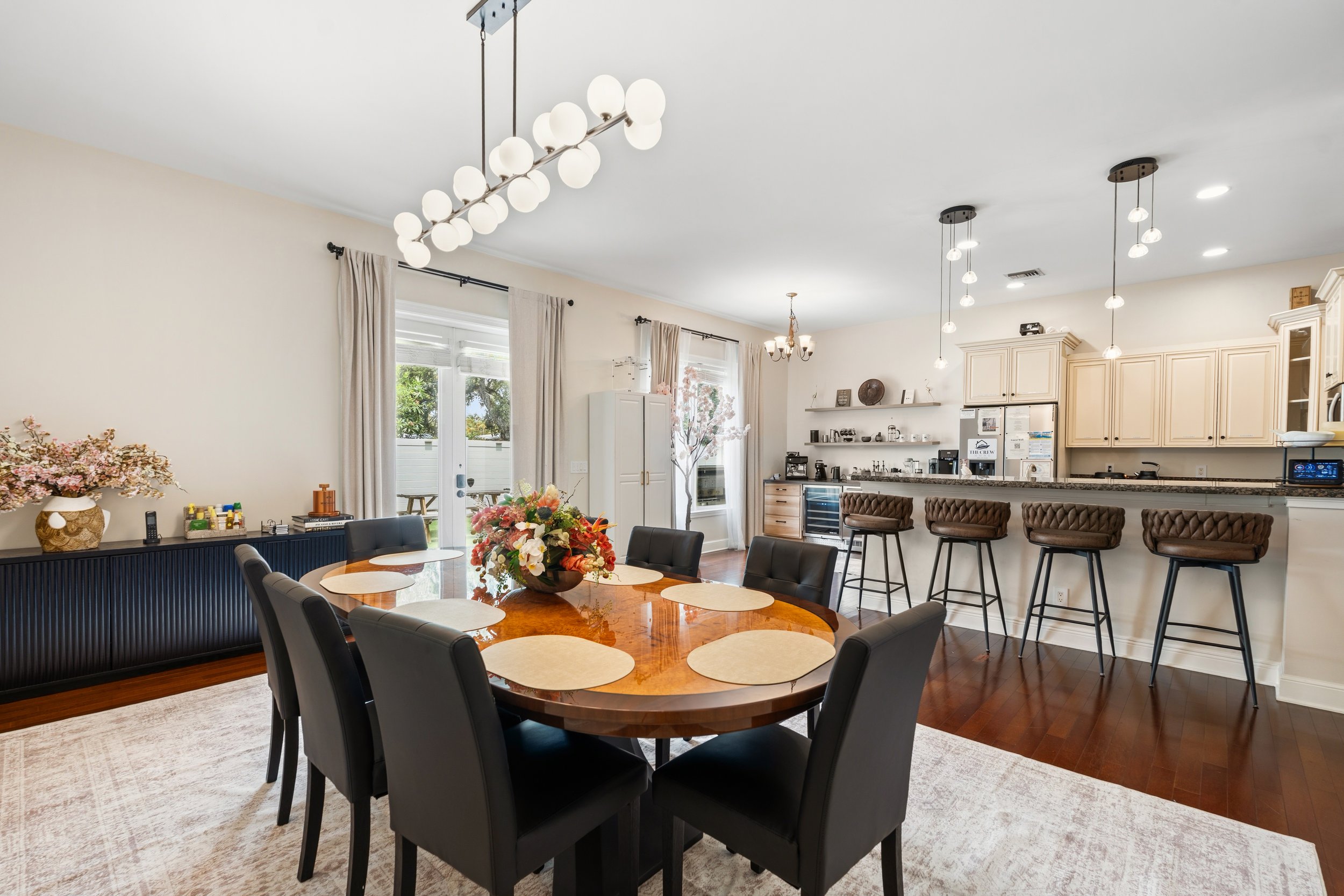 Open-concept kitchen and dining room with a wooden dining table, black chairs, a floral centerpiece, pendant lights, white kitchen cabinets, and bar seating with brown tufted chairs.