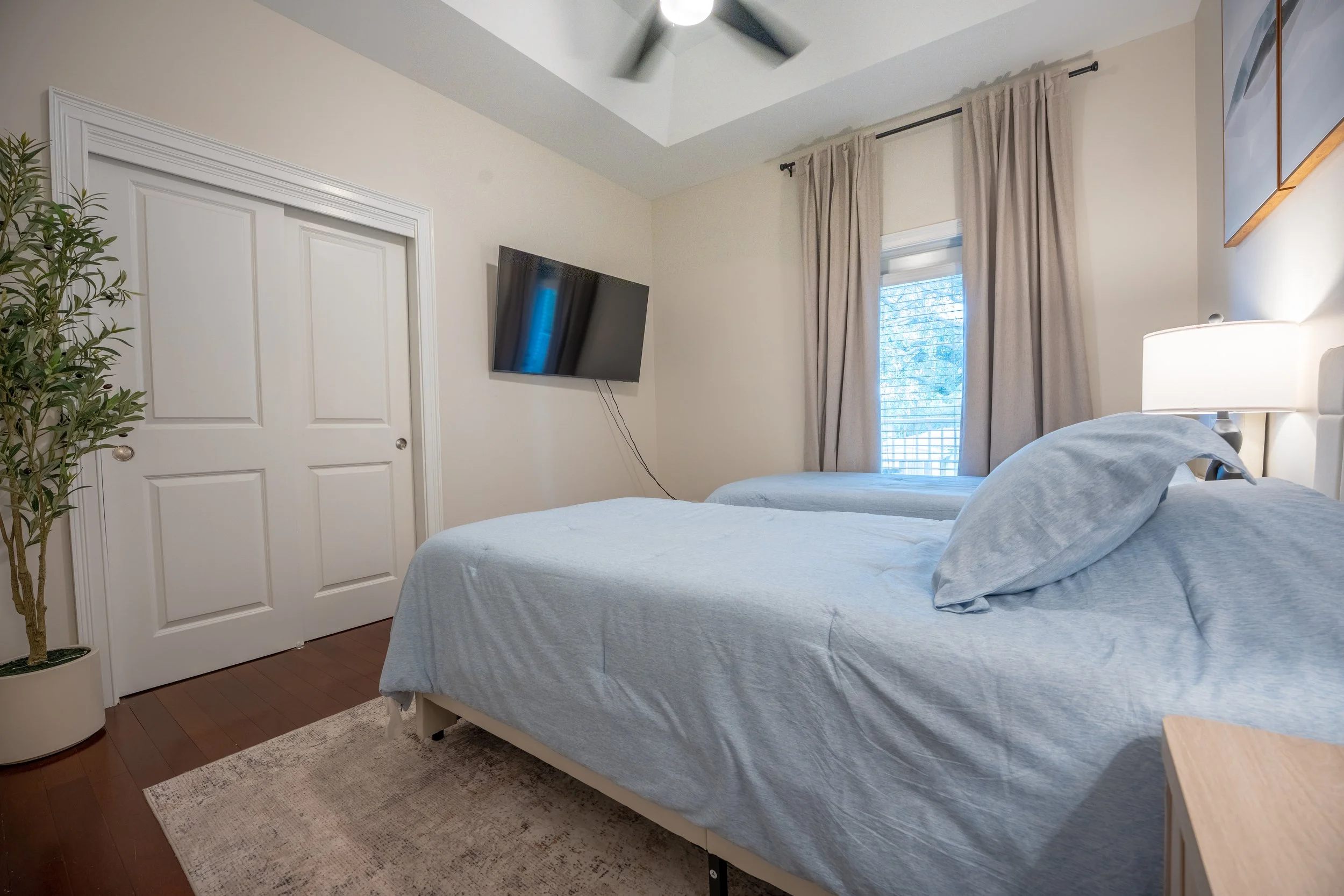 The image shows a bedroom with two beds covered in light blue linens, a flat-screen TV mounted on the wall, a window with beige curtains, a nightstand with a lamp, a potted plant, a door, and a ceiling fan.