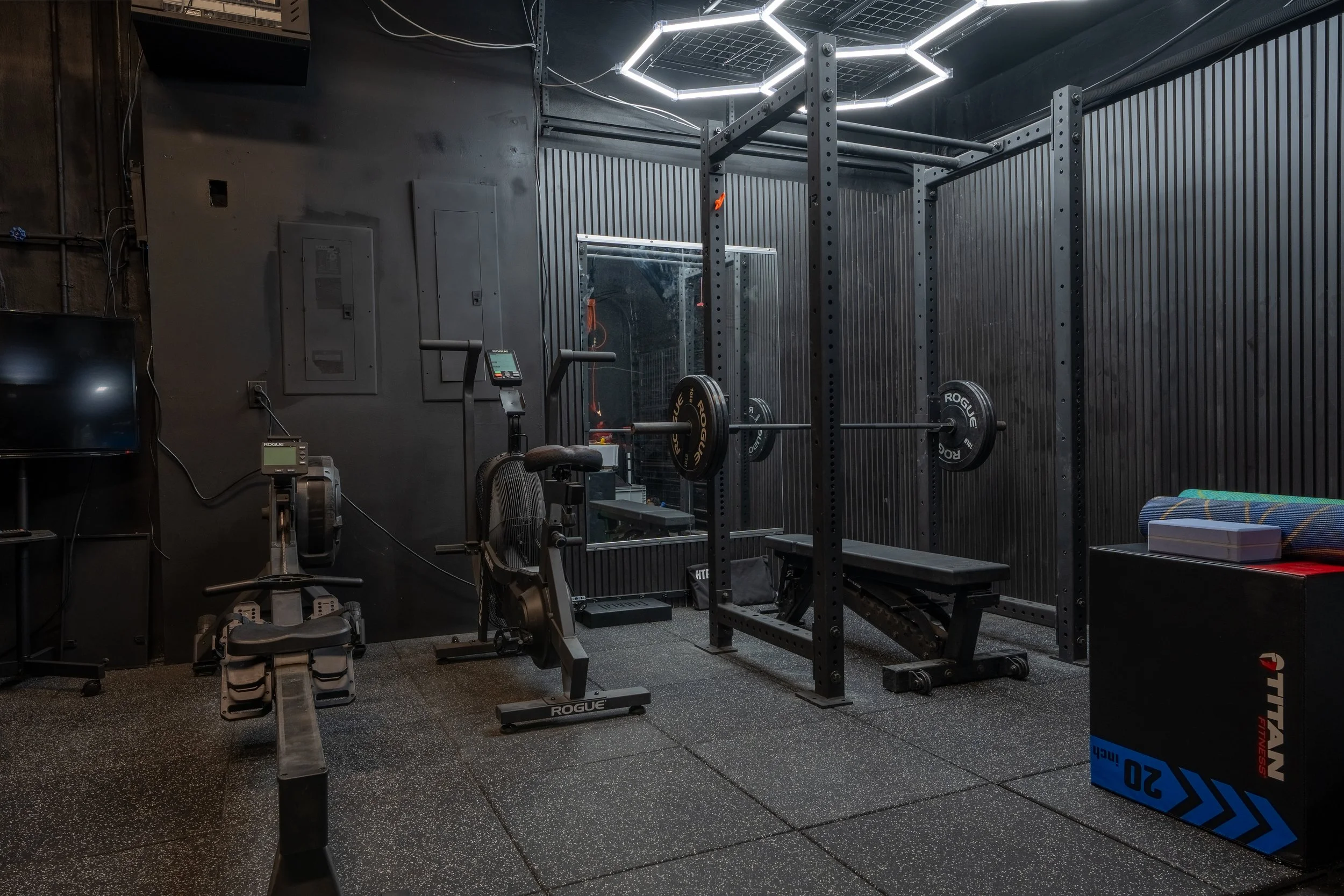 A gym with black walls and flooring, featuring various workout equipment including a squat rack, a rowing machine, a TV, and a blue box. The ceiling has modern geometric lighting.