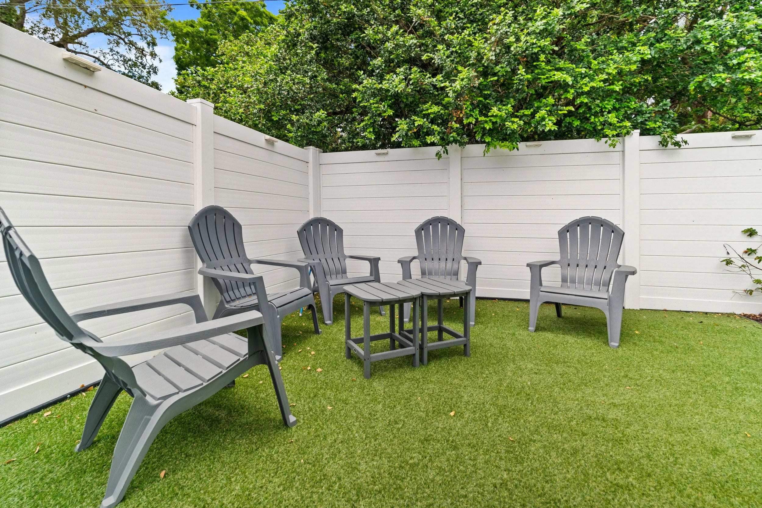 A backyard patio with four gray Adirondack chairs arranged in a semicircle around a small matching table, enclosed by white privacy fencing, with green grass and leafy trees in the background.