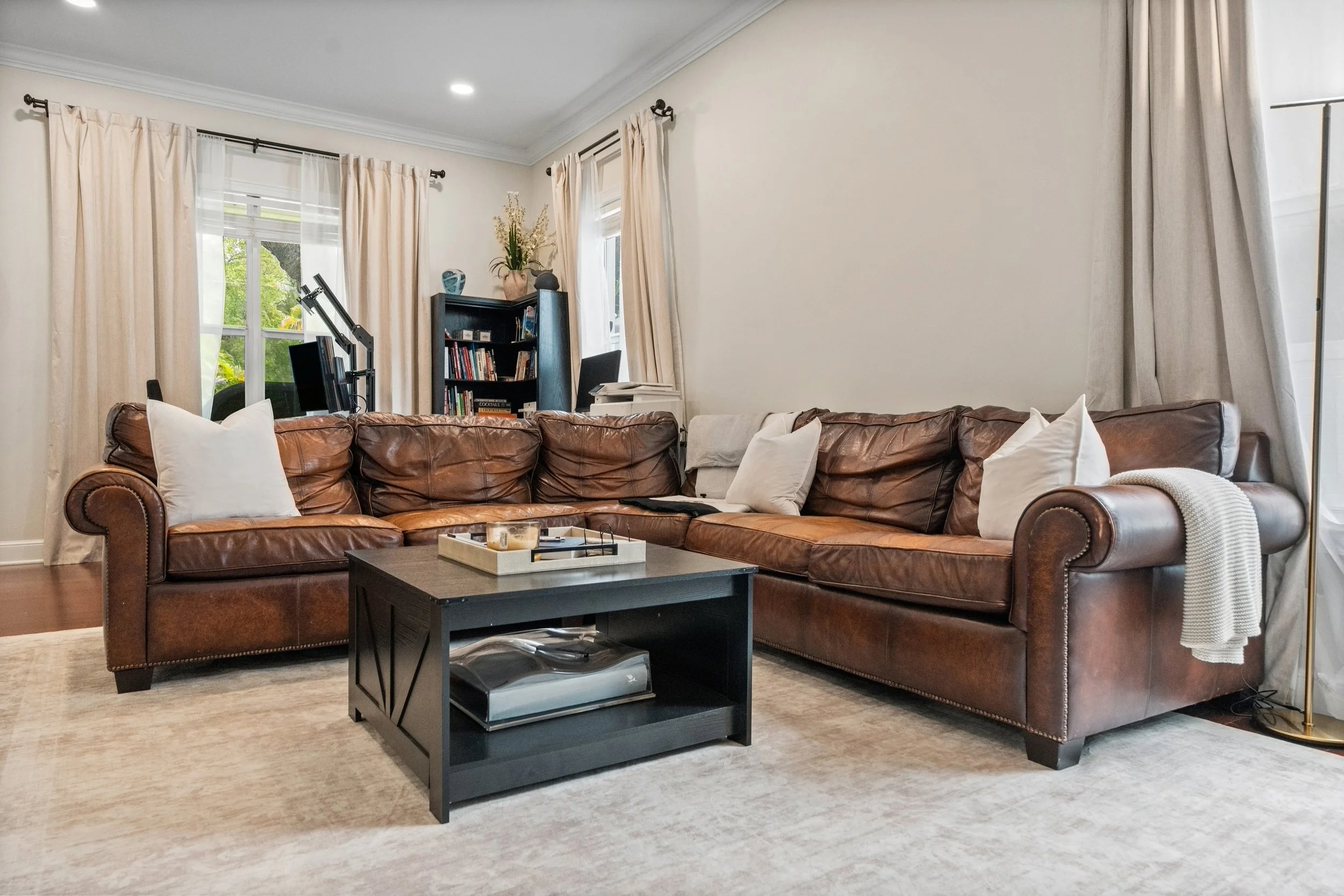 Living room with a large brown leather sectional sofa adorned with white pillows. In front, there is a black coffee table with a tray holding candles. The room has beige walls, four large windows with white and beige curtains, and a black bookshelf f