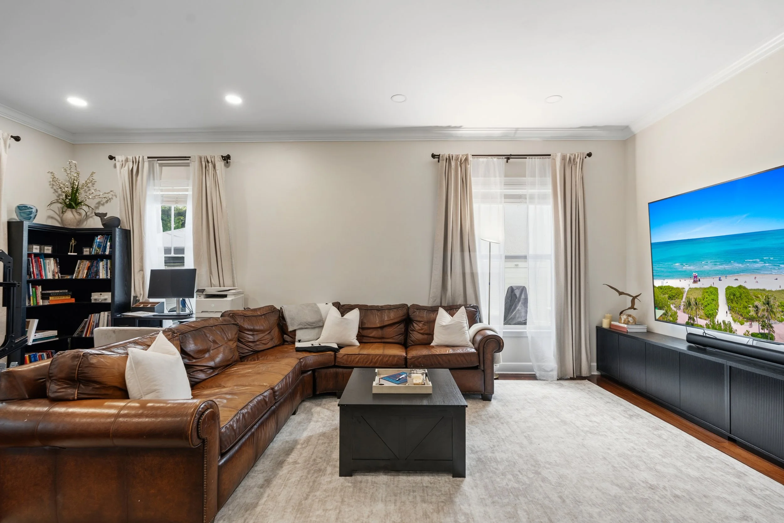 Living room with beige walls, large windows with white curtains, a brown leather sectional sofa with white pillows, a black coffee table with books and a tray, a large flat-screen TV showing a beach scene, a black bookshelf with books and decorative 