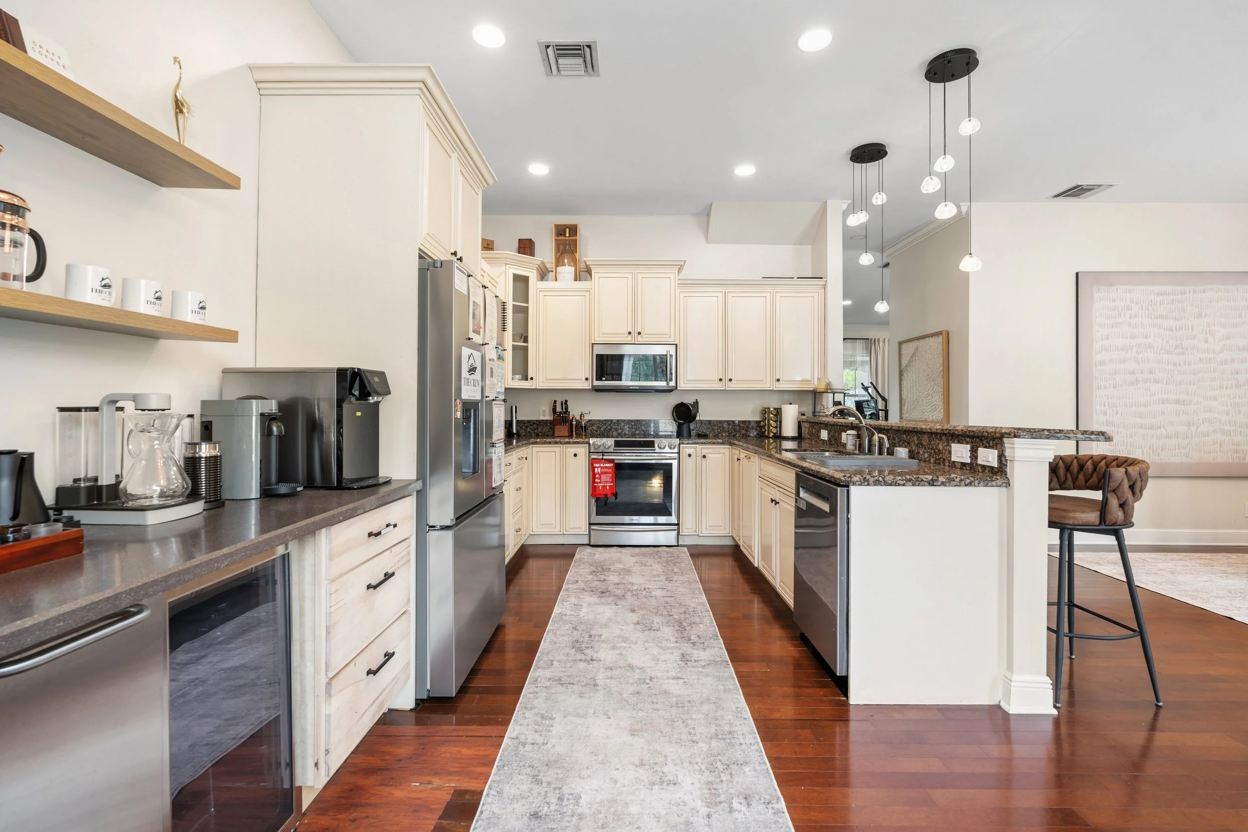 Bright kitchen with white cabinets, stainless steel appliances, granite countertops, hardwood floors, pendant lights, and a gray runner rug.