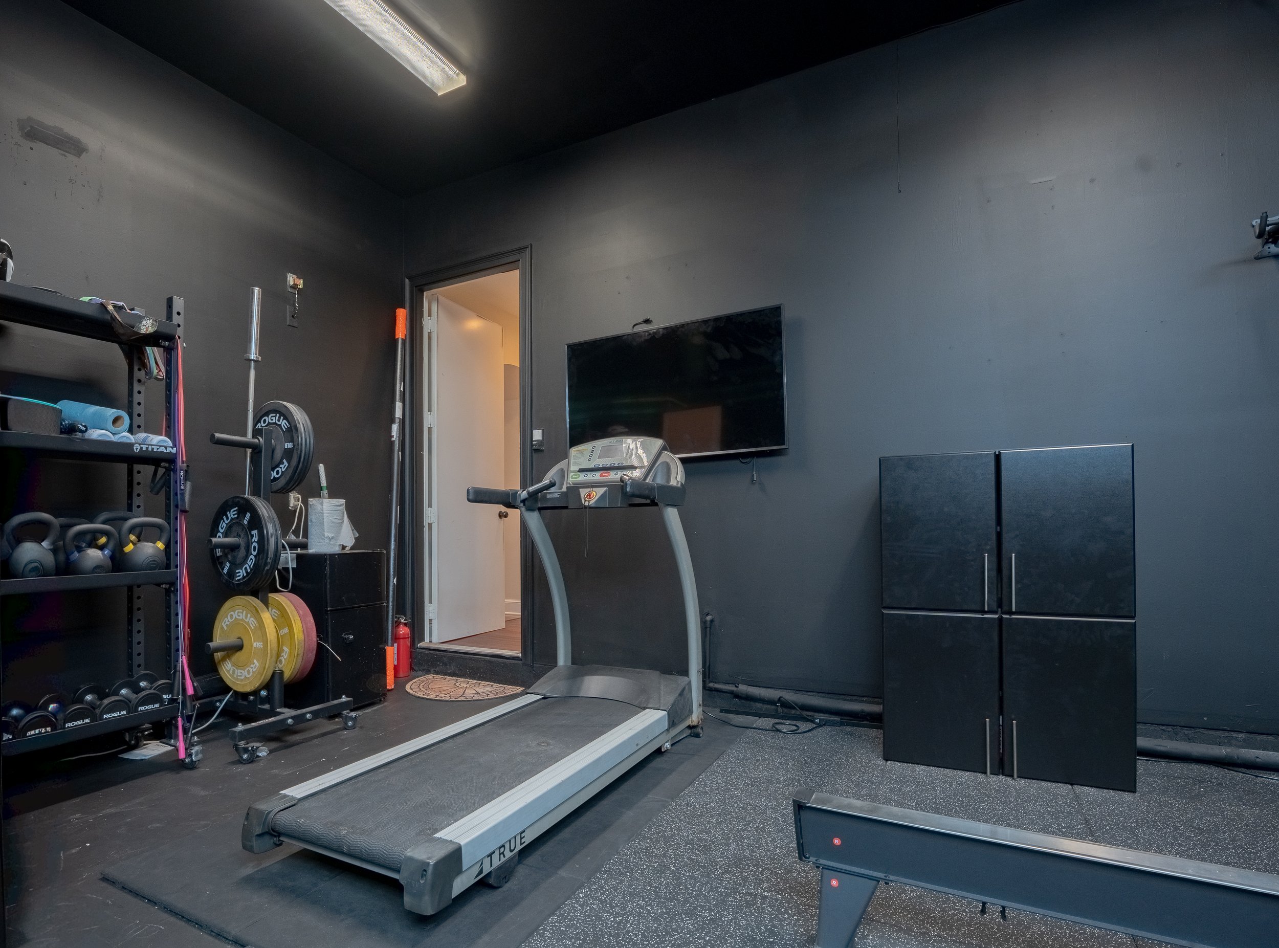 A fitness room with gym equipment, including a treadmill, weights, and a wall-mounted TV, with dark walls and flooring.