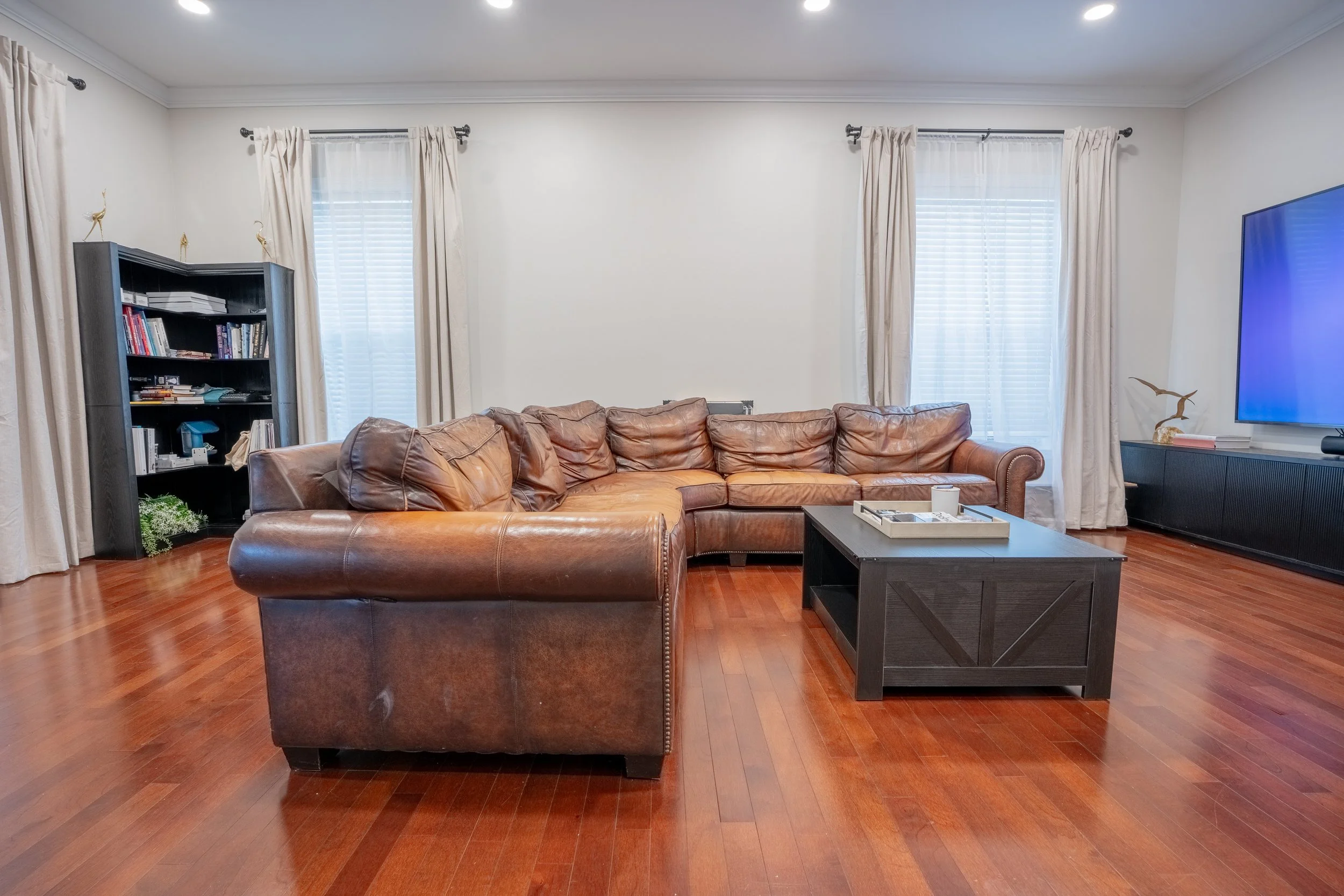 Living room with large brown leather sofa, wooden coffee table, black bookshelf filled with books, and a wall-mounted flat-screen TV, with two large windows with white blinds and beige curtains, wooden flooring, and white walls.