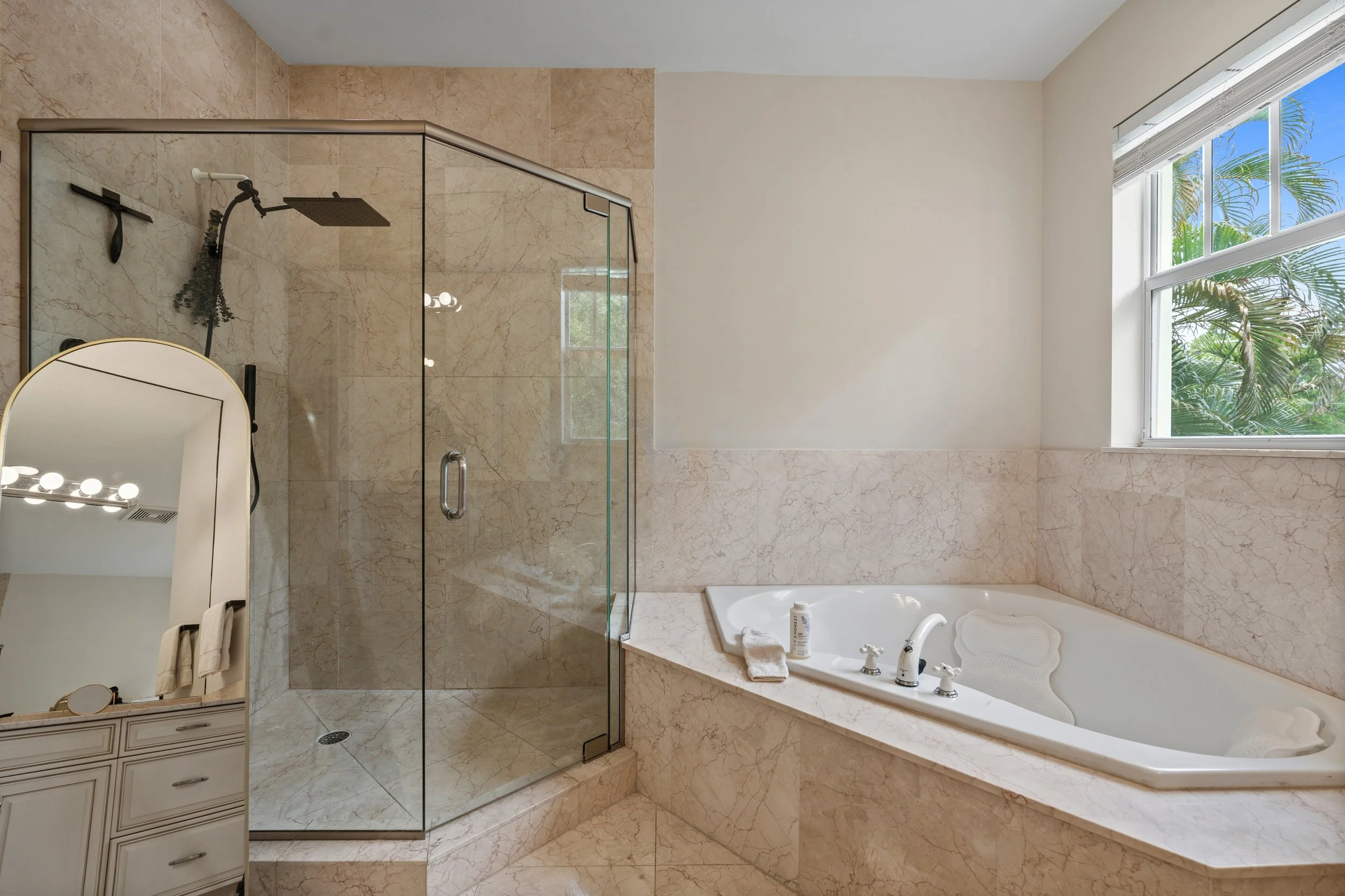 A bathroom with a corner shower with glass doors, a bathtub with a window above, and beige marble walls and floor.