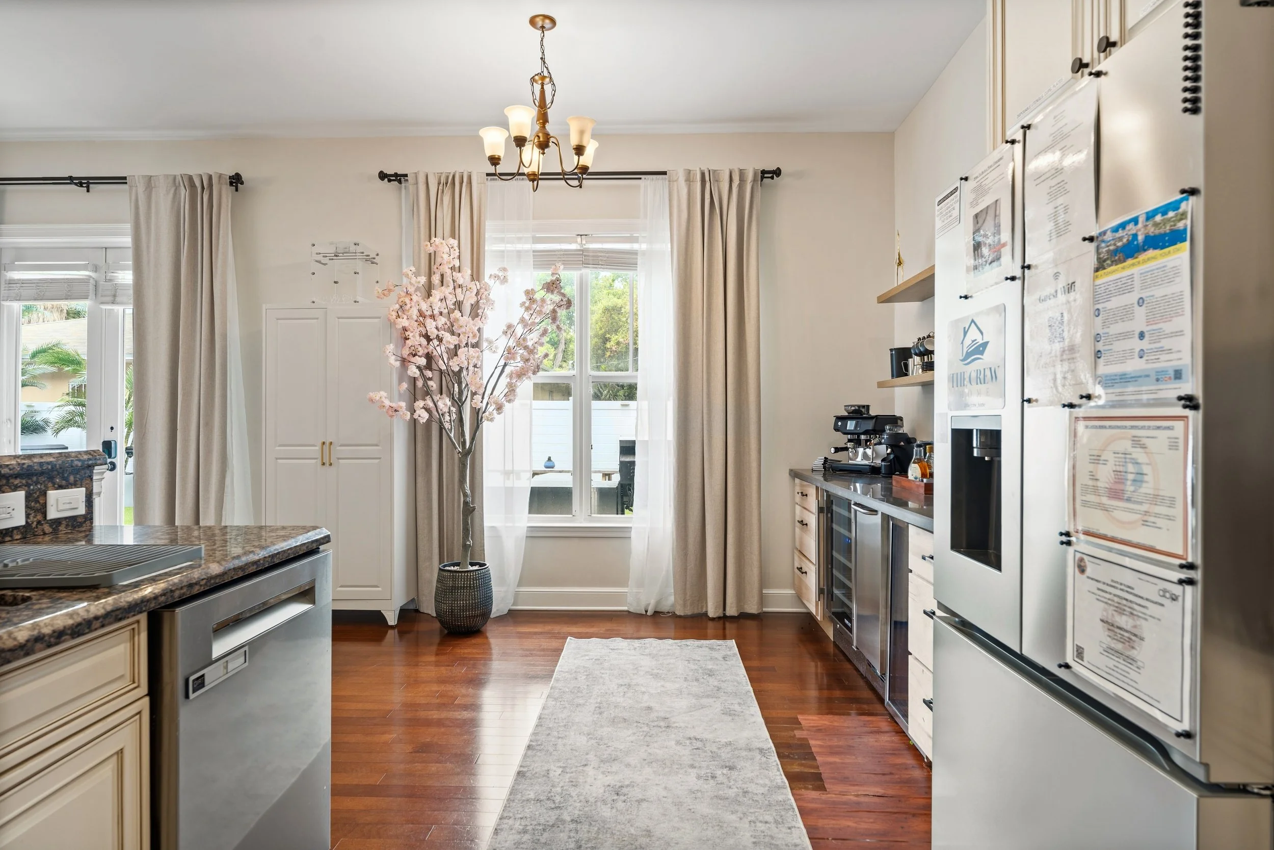 Kitchen with wooden flooring, beige cabinetry, granite countertop, white walls, large window with curtains, a flowering tree in a vase, chandelier, and appliances including a refrigerator and coffee machine.