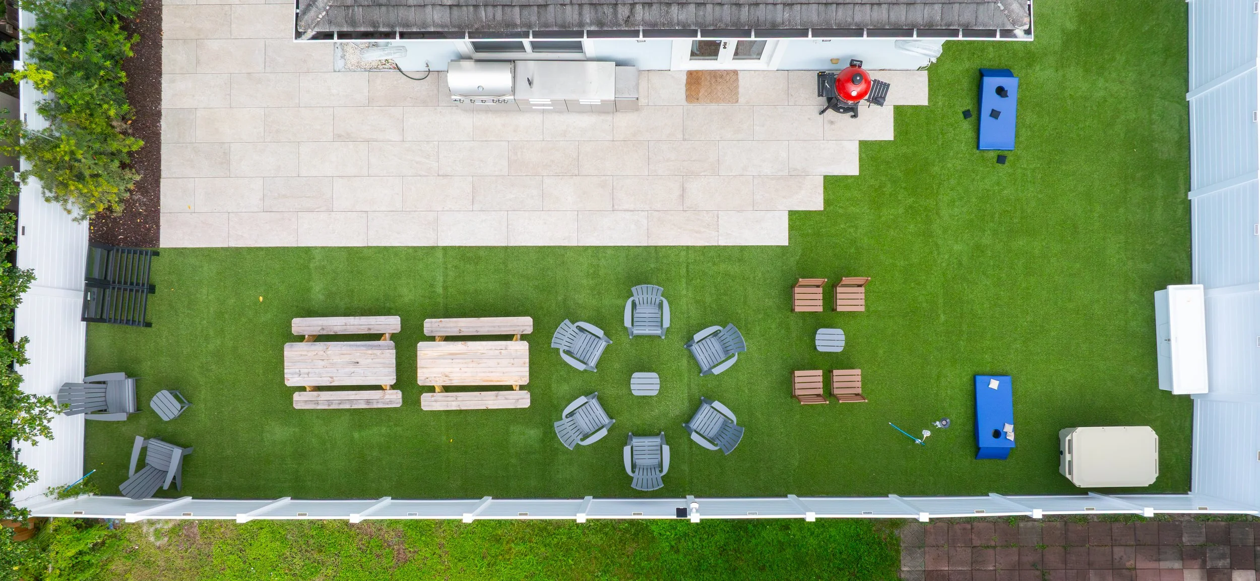Top-down view of a backyard with a lawn, patio area, outdoor furniture including chairs and tables, a grill, and various outdoor equipment.