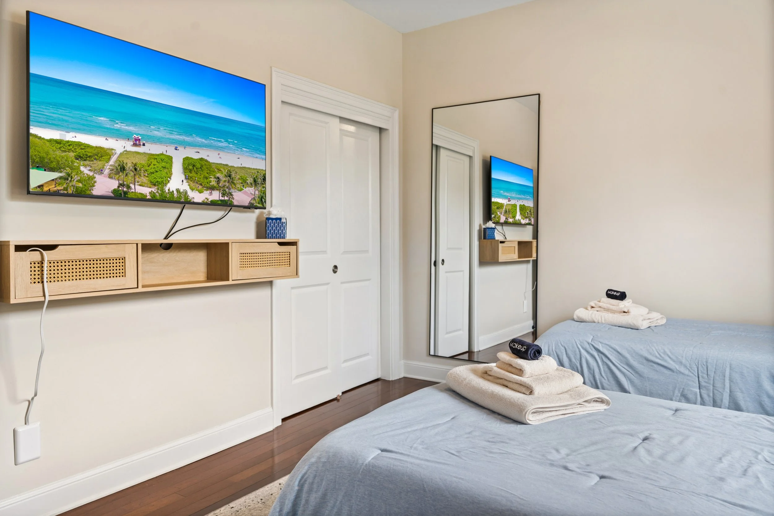 A bedroom with two twin beds covered with light blue sheets, with neatly folded white towels and dark gray rolled towels labeled 'Makeup' on top. There is a large mirror on the wall, a sliding closet door, a wall-mounted TV showing a beach scene, and