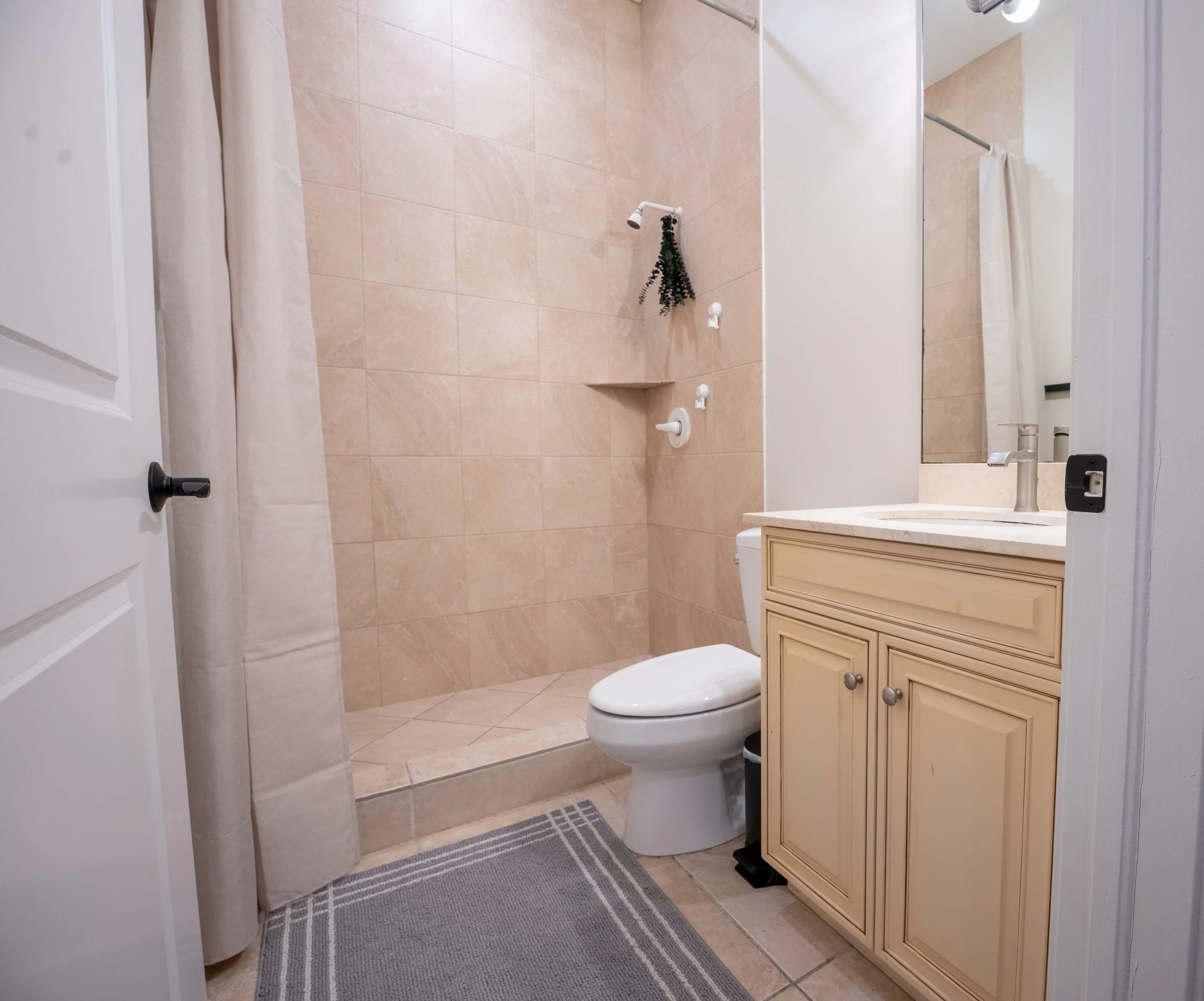 Bathroom with beige tiled walls, a white toilet, a cream-colored bathroom vanity with a mirror, a sink, and a grey striped rug on the floor. There is a shower curtain on the left side and a small shelf in the shower area holding a green plant decorat