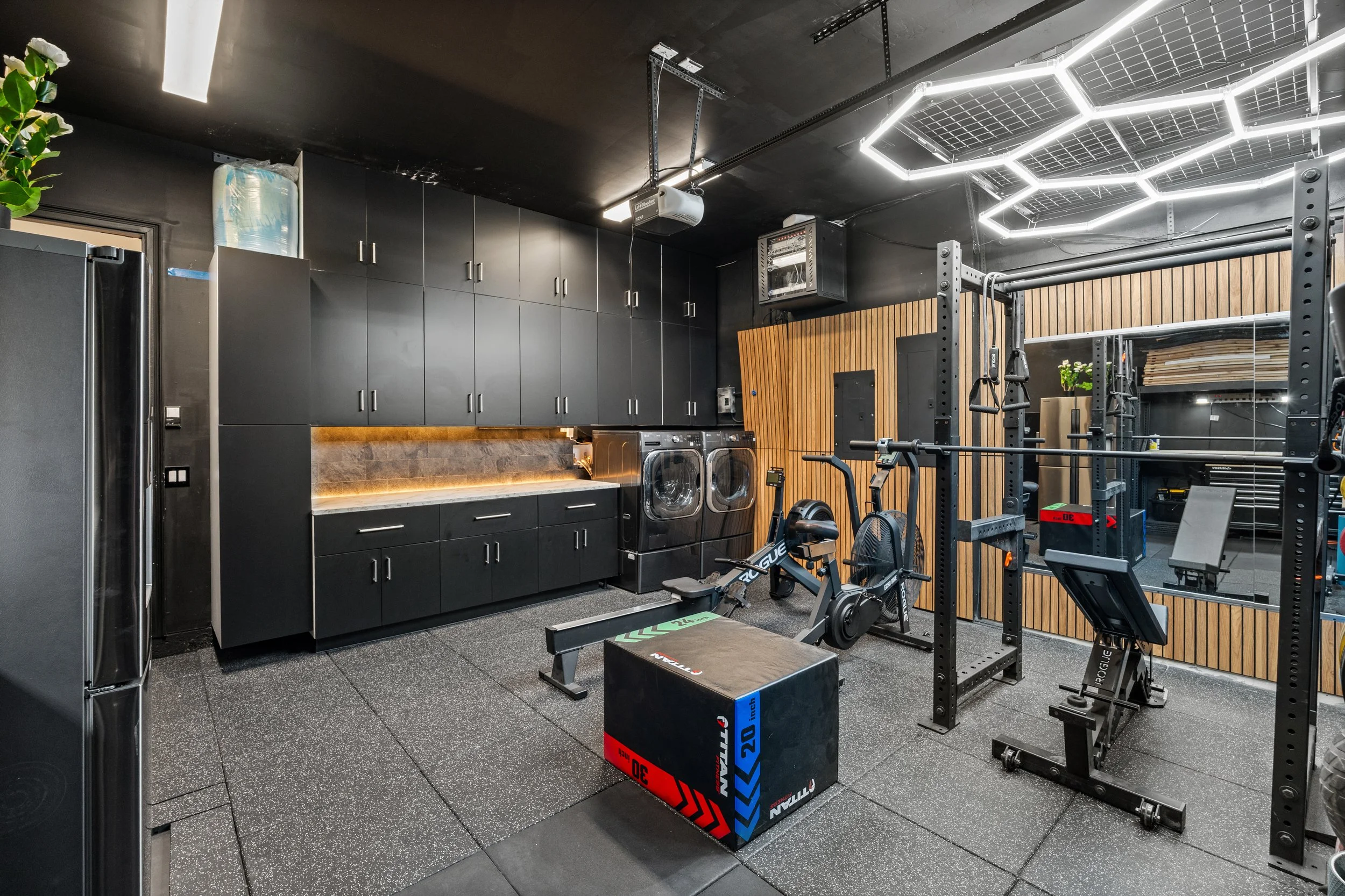 Interior view of a modern gym with black and wooden decor, featuring workout equipment such as a rowing machine, pull-up bars, medicine balls, and black cabinets.