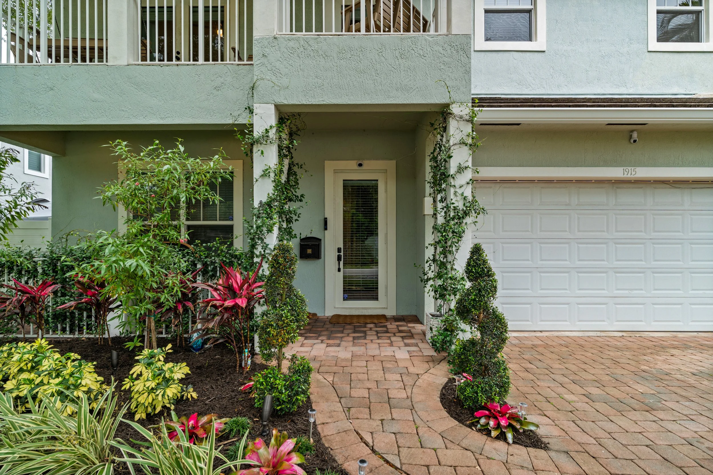Front view of a house with a garden and brick pathway. The house is painted light green with white trim, featuring a front door with a glass panel and a security camera. There is a white garage door to the right, and the garden in front has various g