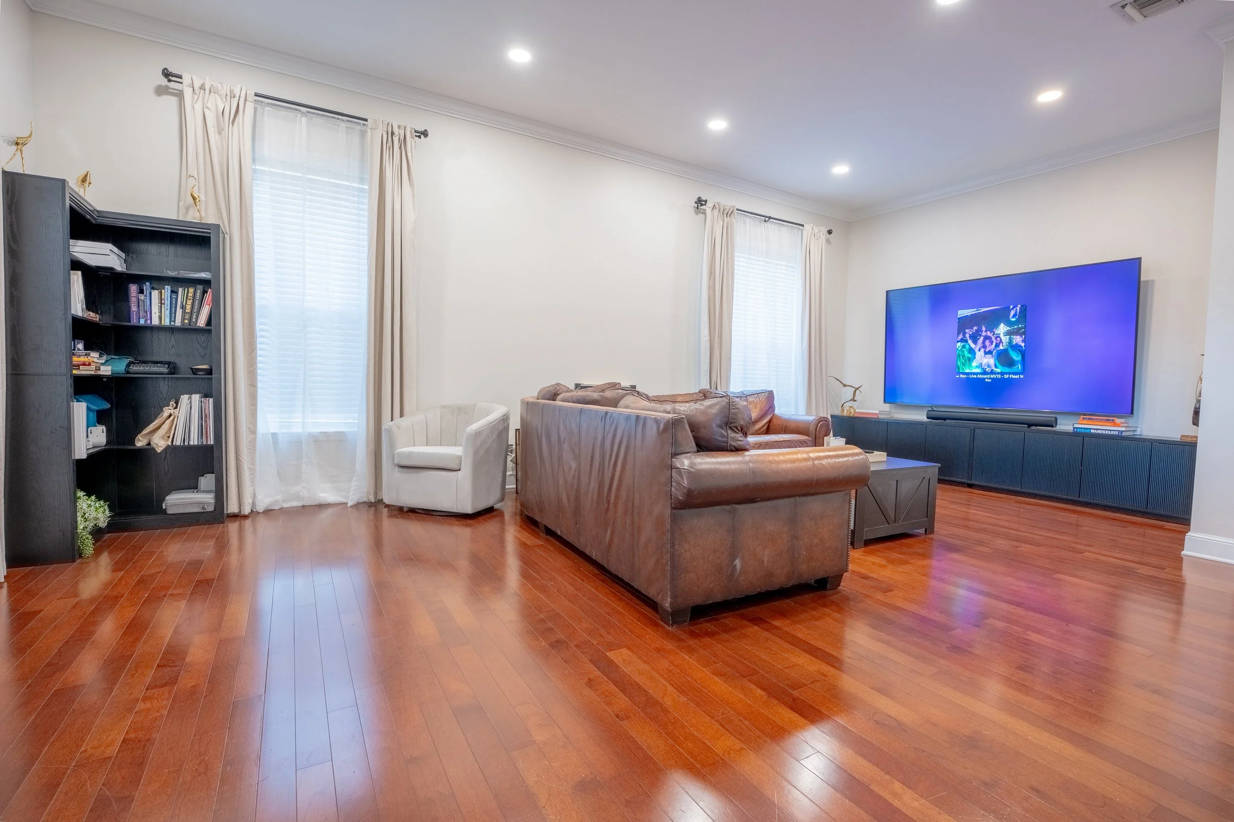 Living room with hardwood floors, white walls, and windows with curtains. Contains a black bookshelf with books and decorative items, a white armchair, a brown leather sofa, and a large flat-screen TV on a black TV stand. The room is well-lit with re