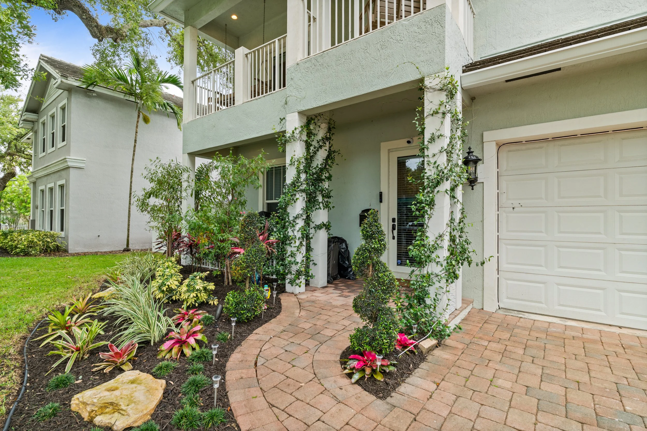 Front view of a light green house with a brick pathway, lush plants, flowers, and vines growing around the entrance, with trees and a clear sky in the background.