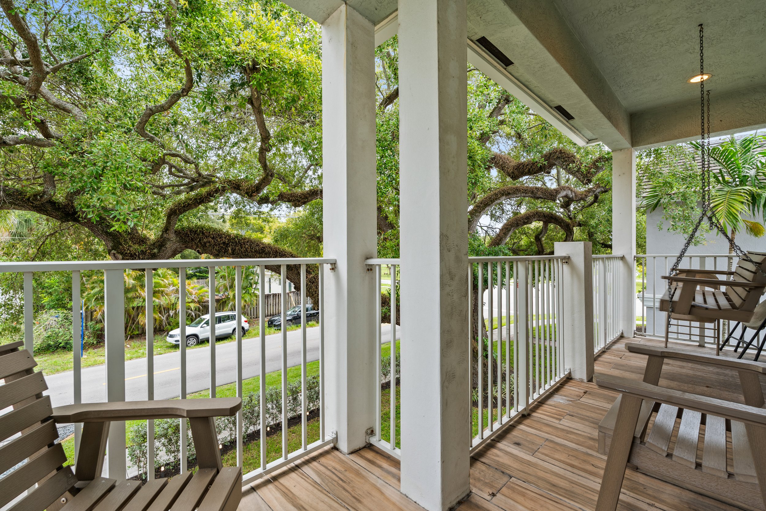 View from a porch with wooden flooring, white railing, and white columns, overlooking a street with parked cars, large green trees, and a garden.
