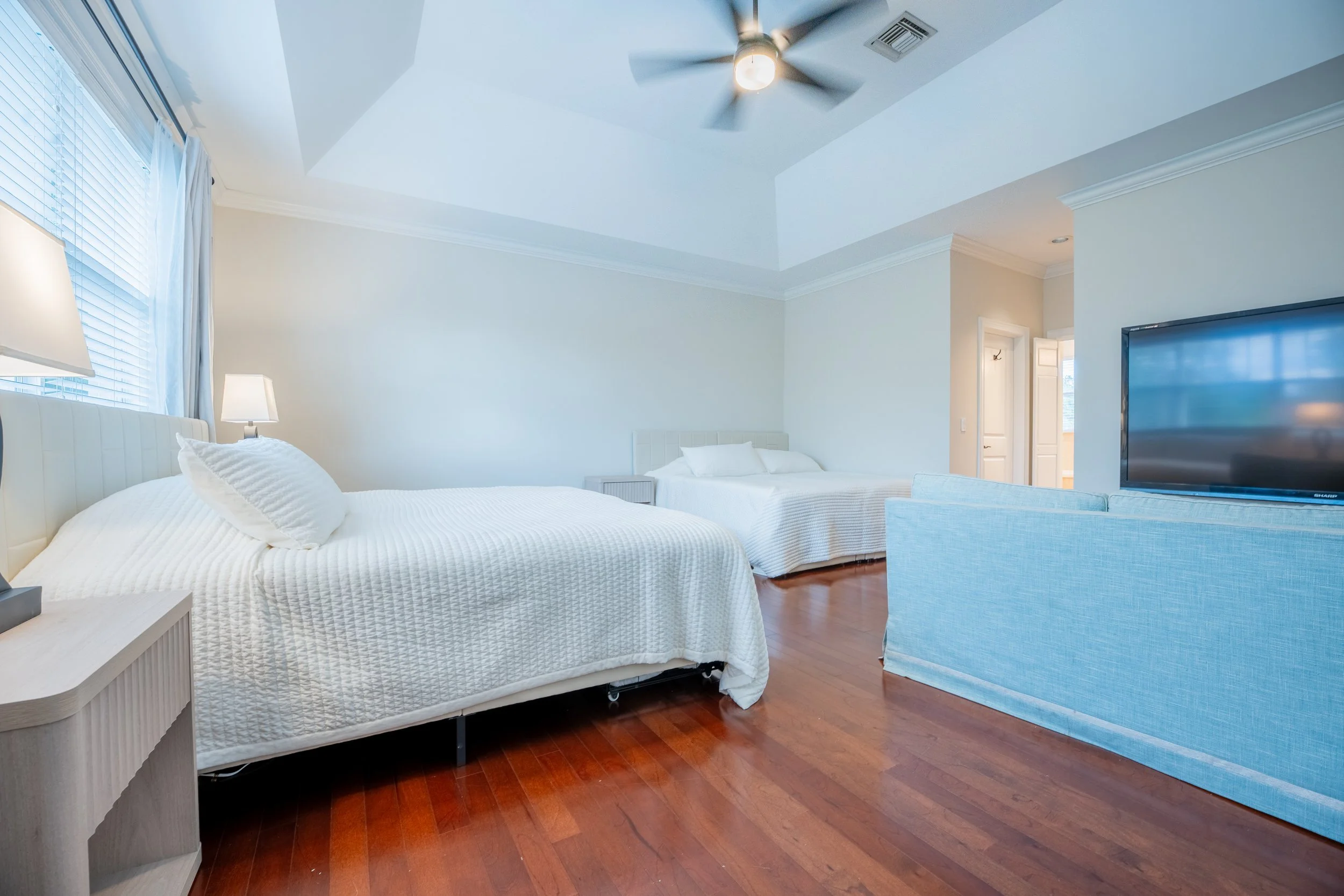 A bedroom with two white beds, a flat-screen TV, wooden floor, and a ceiling fan.