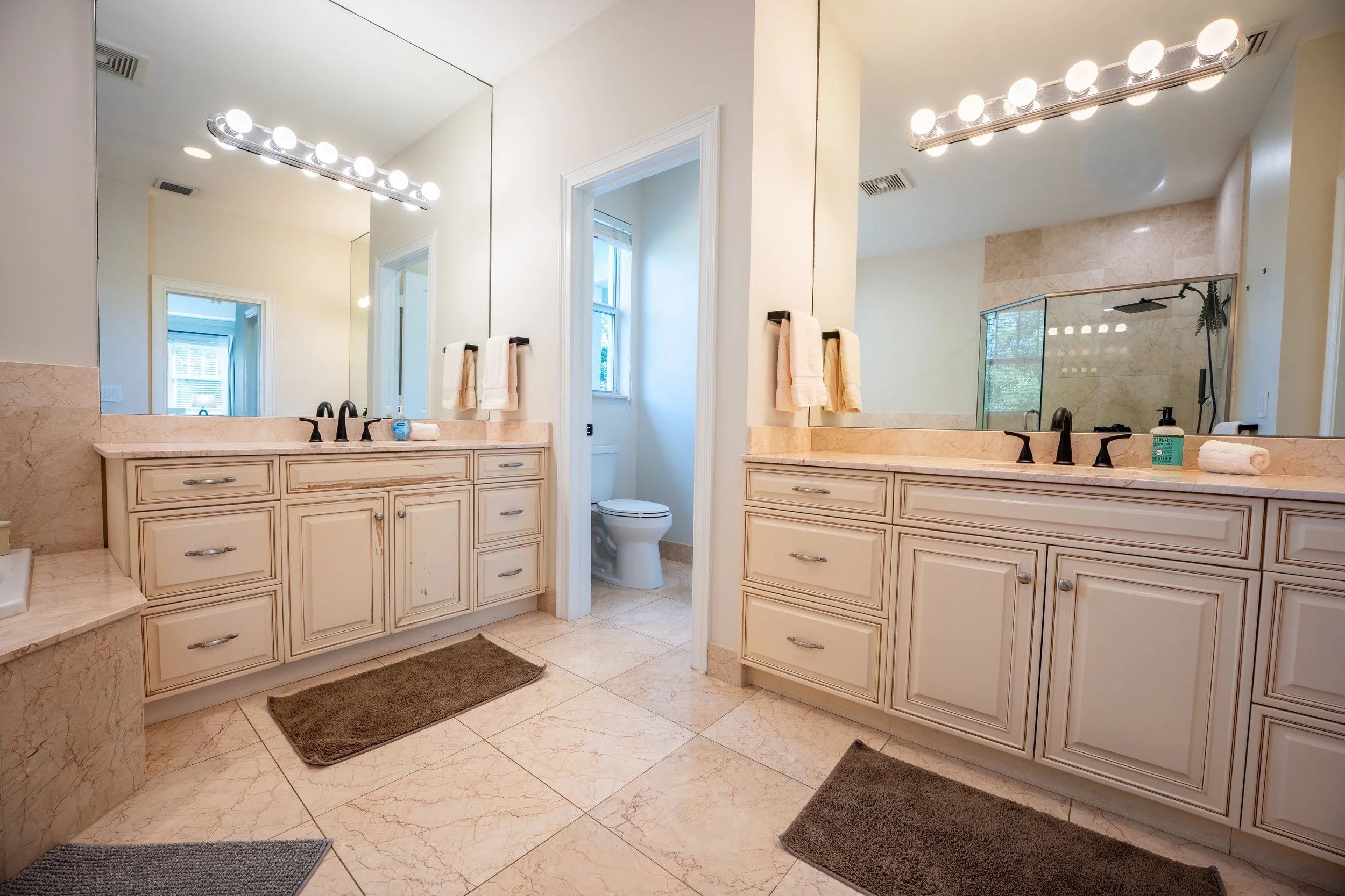 A spacious bathroom with dual vanities and large mirrors, beige cabinetry, a toilet in a separate room, and a walk-in shower with a glass door, illuminated by bright lighting.