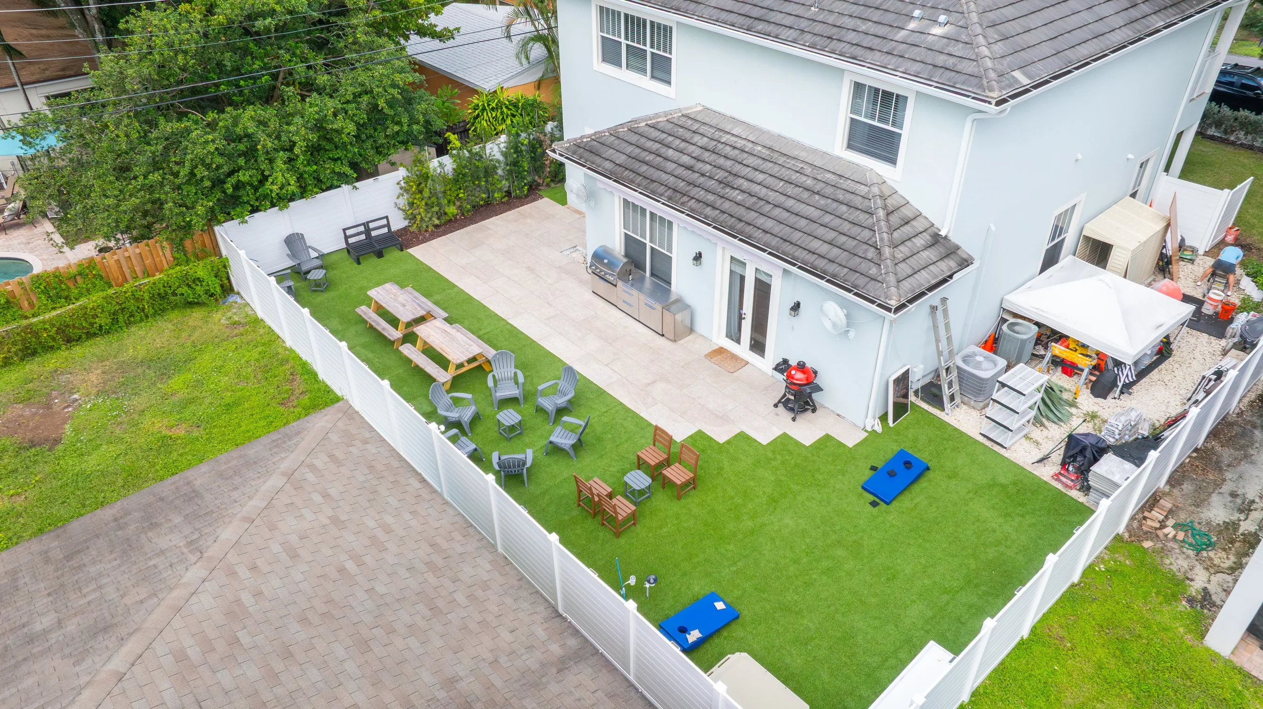 A backyard view from above showing a white fence enclosing a green lawn area with outdoor furniture, a patio with a built-in grill, and a section under construction with tools and equipment near a two-story light blue house with a gray roof.