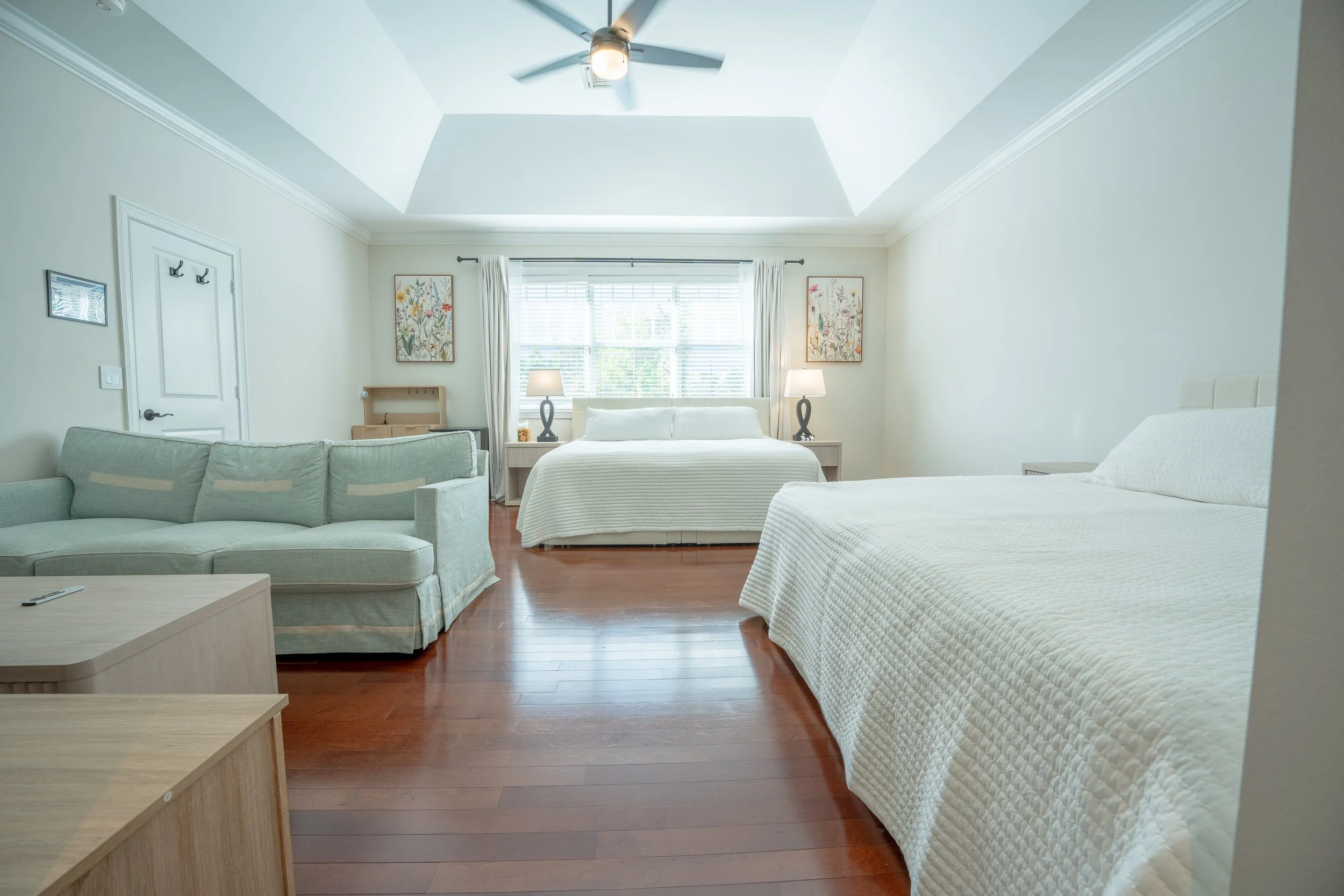 A spacious bedroom with two beds, a sofa, a ceiling fan, a large window with curtains, and two table lamps, decorated with floral artwork.