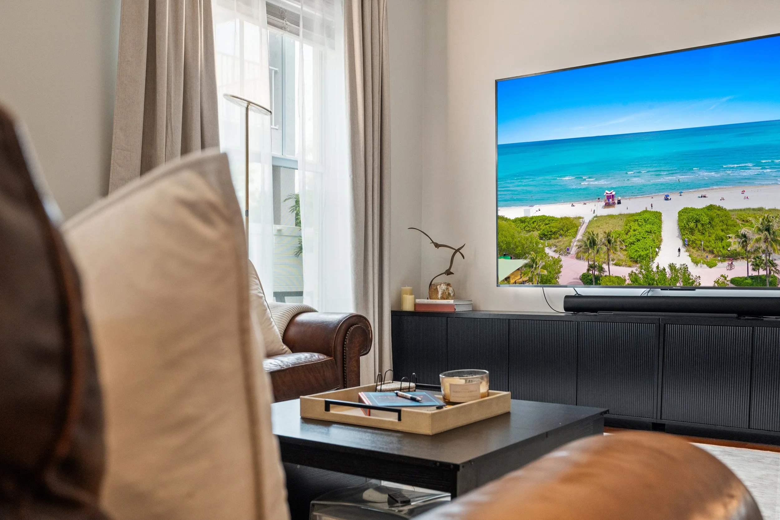Living room with beige curtains, wooden coffee table with tray and books, leather sofa, TV displaying a beach scene with sand, green bushes, and blue ocean.