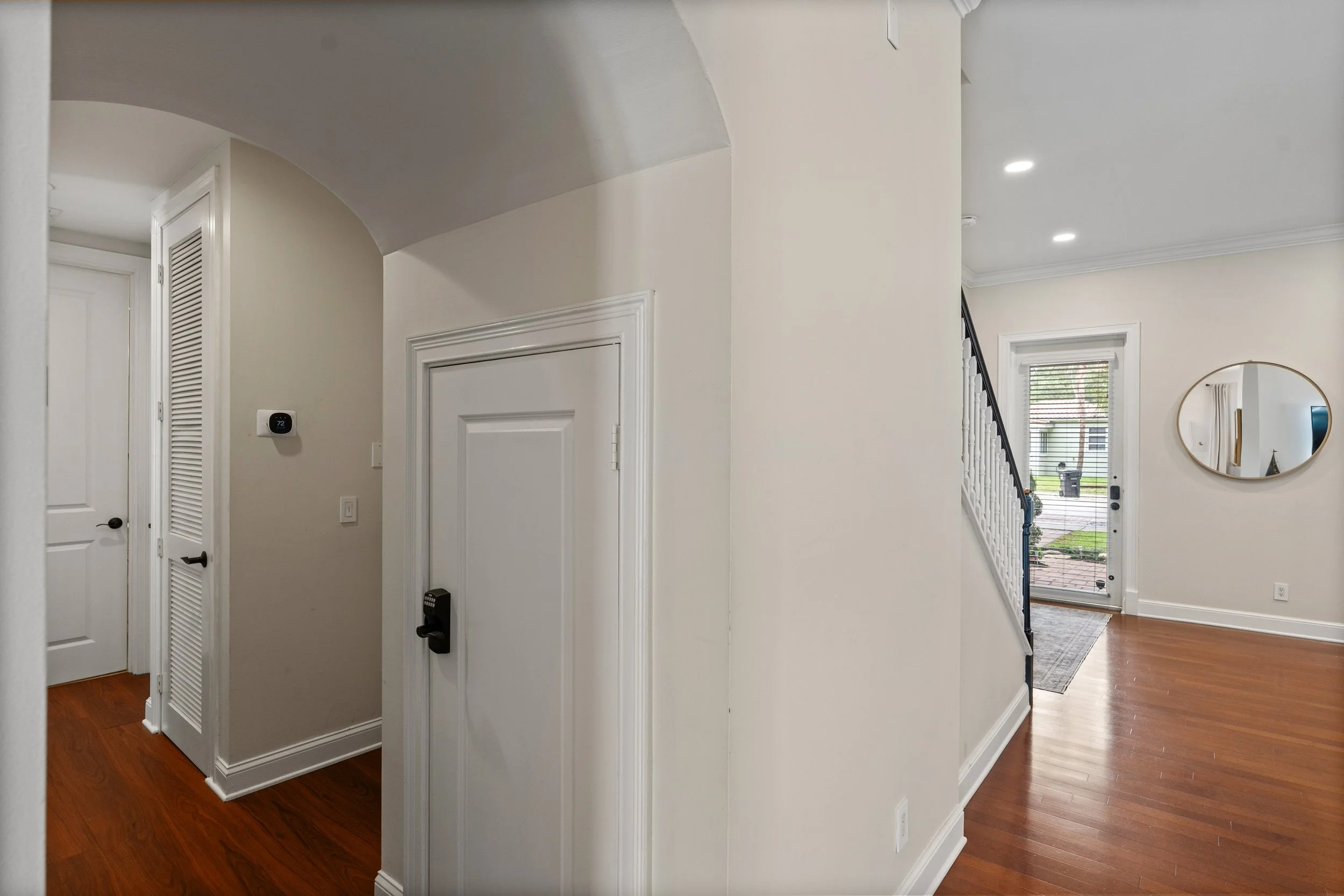 Interior view of a house's entryway with a white door, a round mirror on the wall, and a staircase leading to the upper floor, with a view of the front door and outside greenery.
