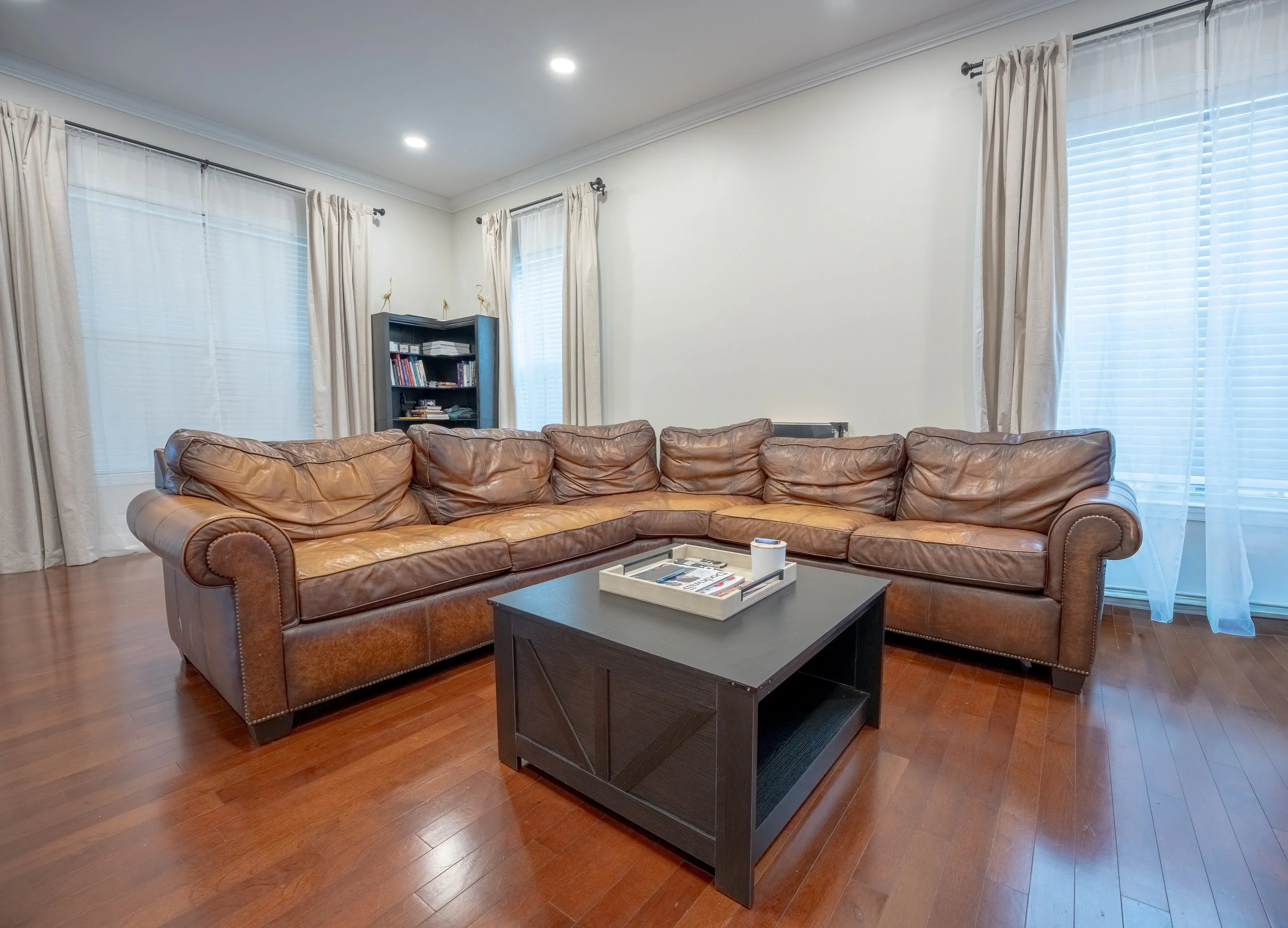 Living room with a brown leather sectional sofa, black coffee table, white tray with magazines, cream curtains on large windows, and a blue bookcase with books and decorative cranes.
