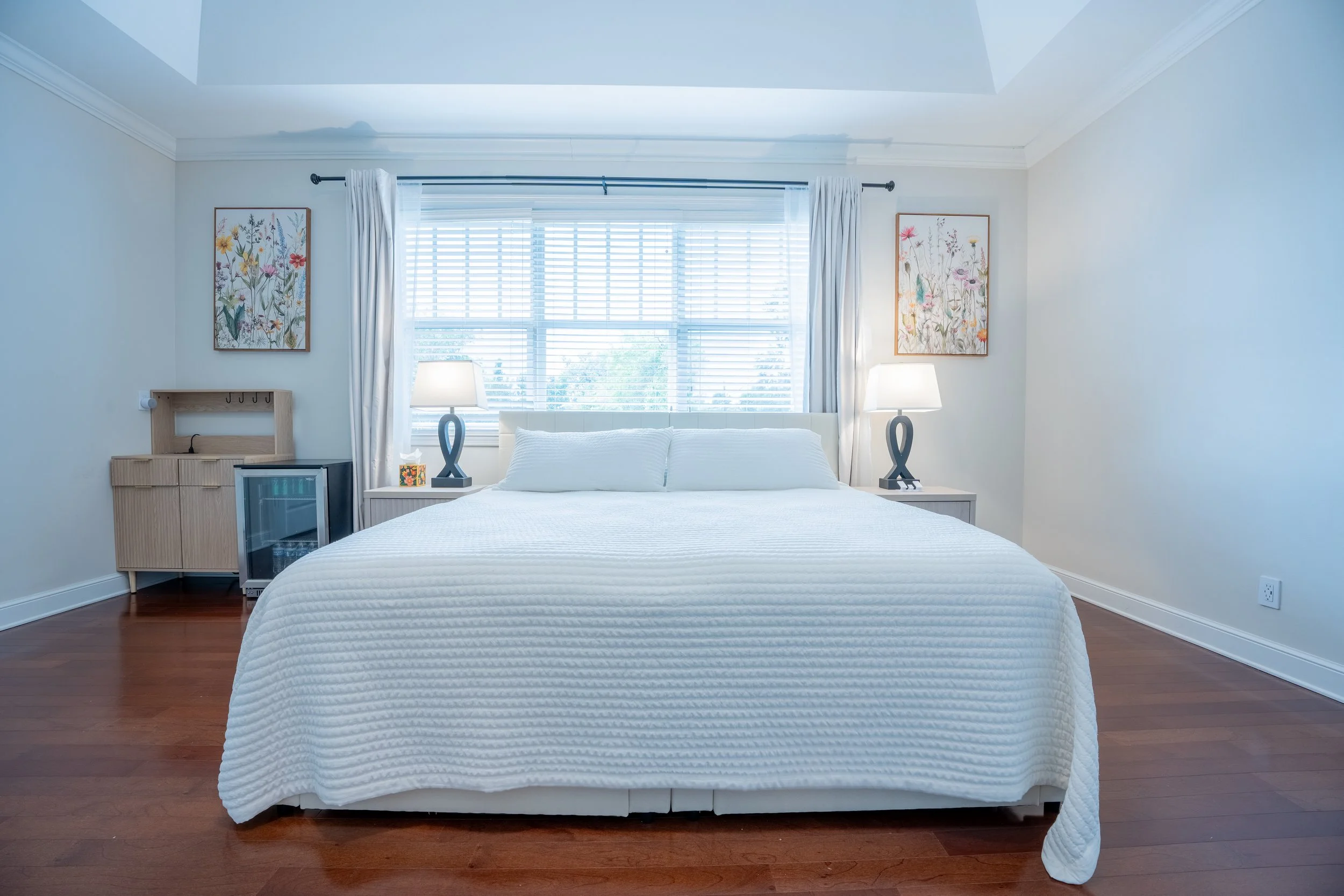 A neatly made bed with white bedding in a bright bedroom. Two bedside tables with lamps are on either side. A window with blinds and white curtains is behind the bed, and artwork with floral themes is hung on the wall.