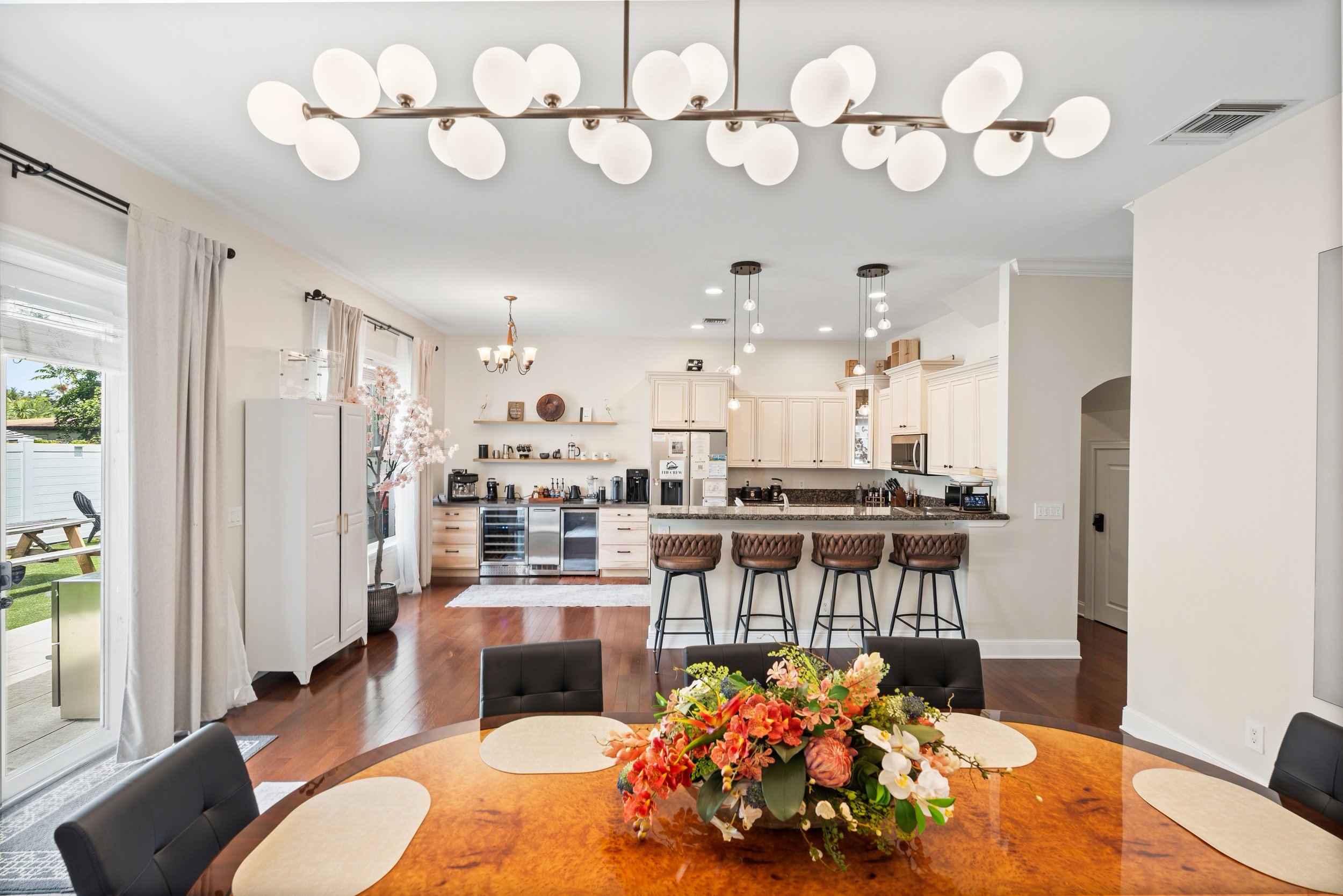 Interior of a modern kitchen and dining area with a wooden dining table featuring a floral centerpiece, black chairs, white cabinetry, multiple pendant and ceiling lights, and sliding glass doors with beige curtains.