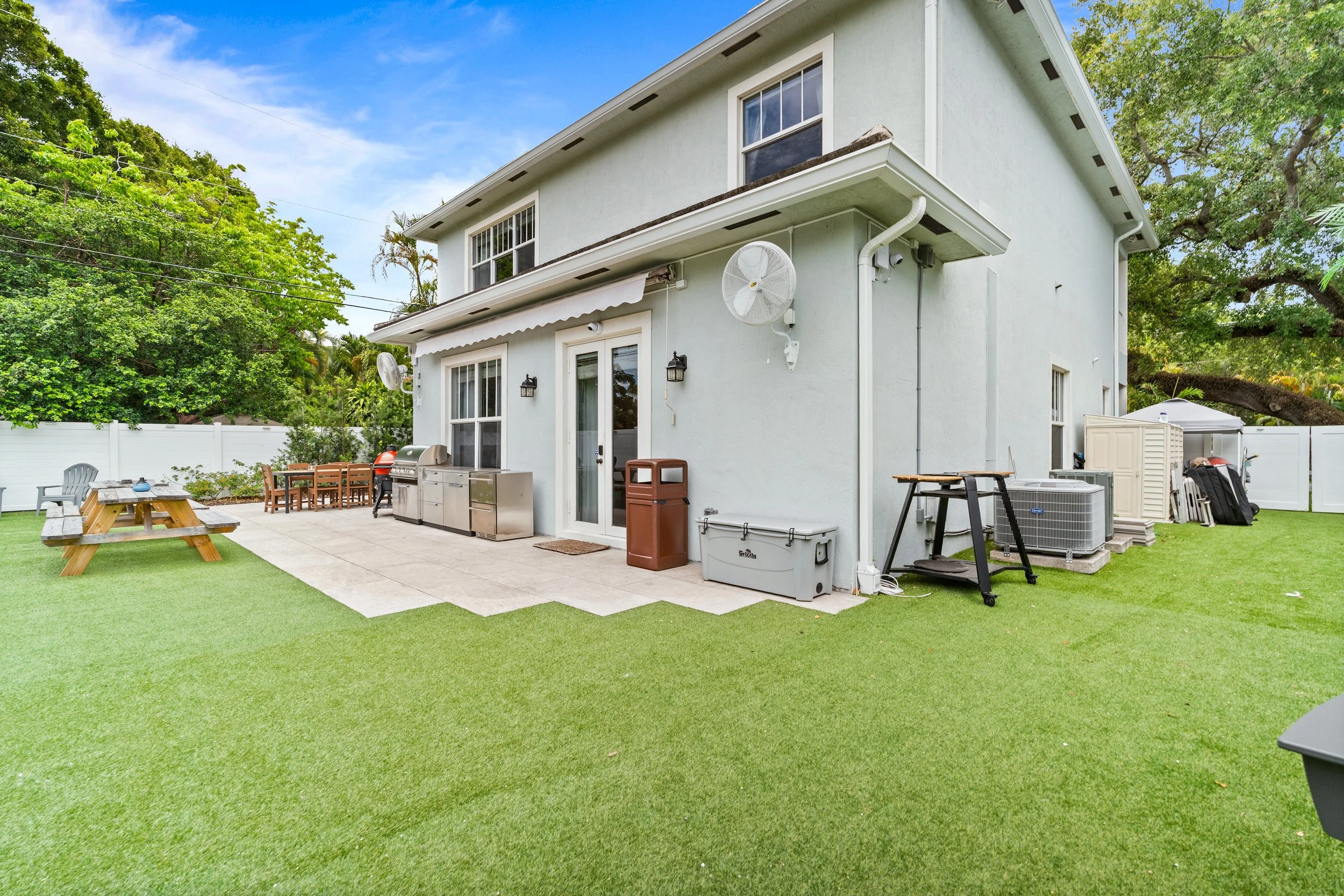 Backyard patio area with outdoor kitchen, tables, chairs, and green artificial grass, adjacent to a light-colored house with large windows.