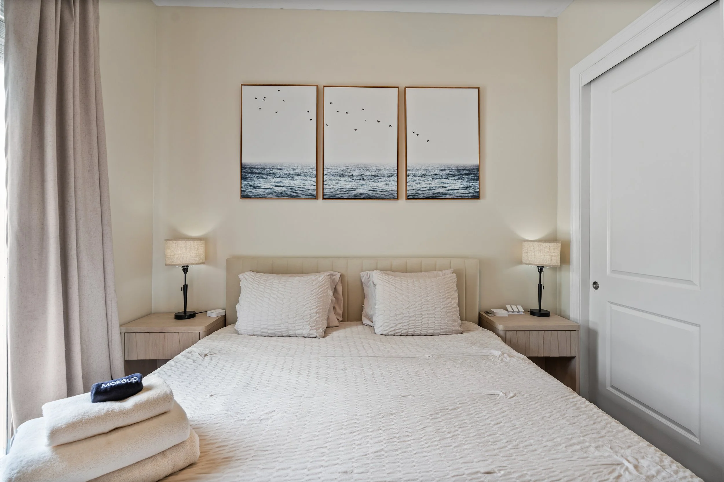 A neatly made bed with white textured bedding and two matching pillows. On top of the bed, there are two folded beige towels with a dark blue makeup towel on top. Two bedside tables with lamps are positioned on either side of the bed. A triptych of a