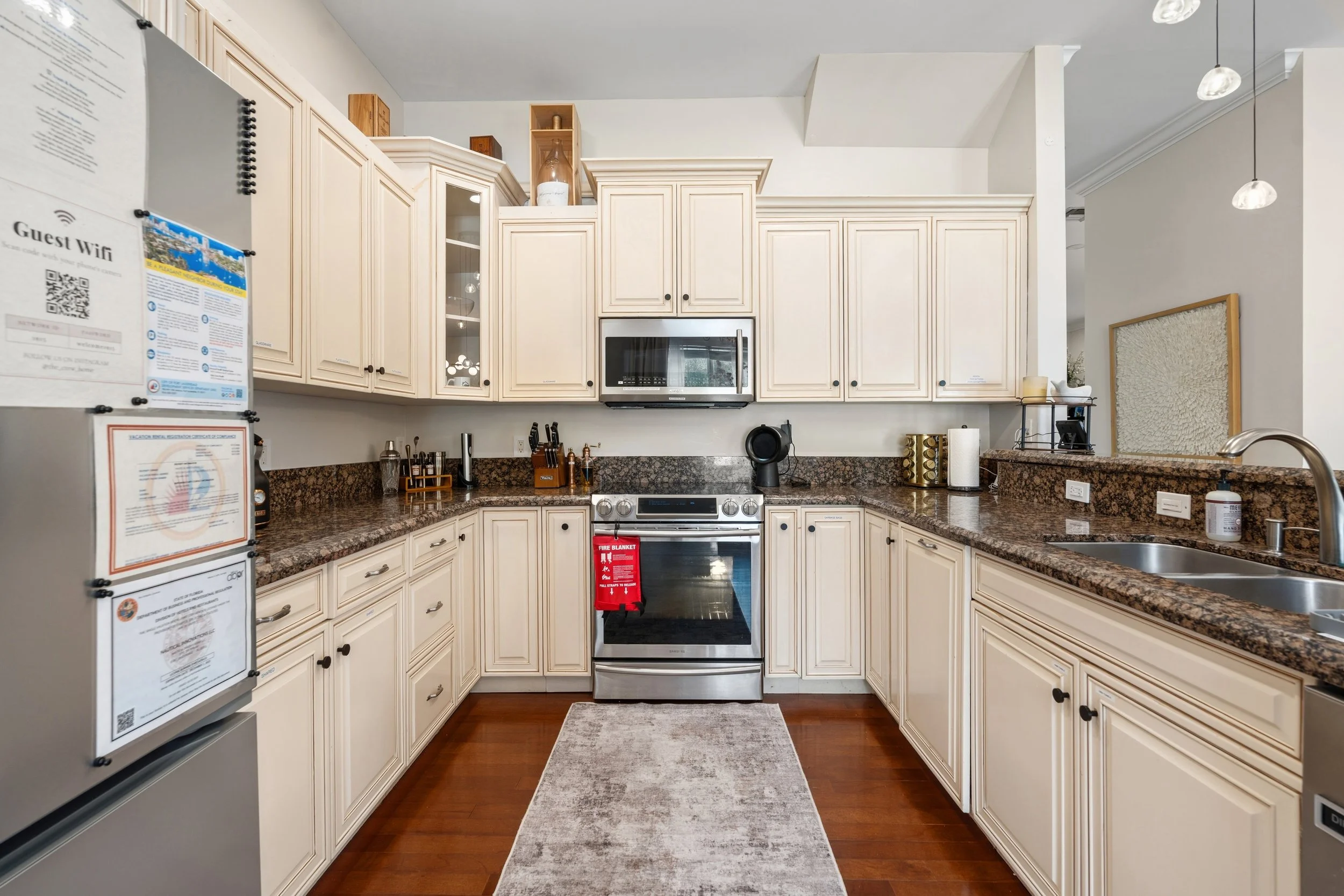 Kitchen with cream-colored cabinets, granite countertops, stainless steel oven and microwave, sink, and various kitchen items. A refrigerator with notices and and a gray rug on the floor.