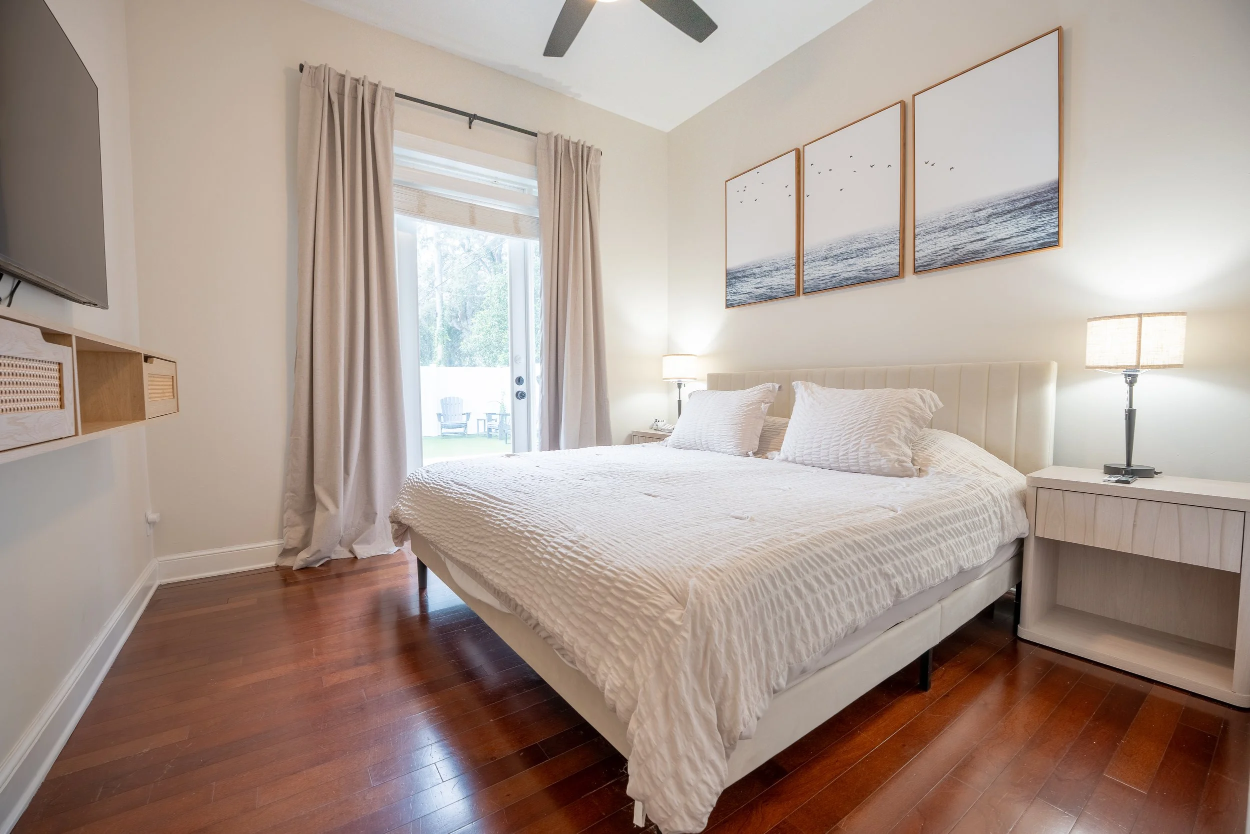 A bedroom with a bed, white bedding, and pillows, a nightstand with a lamp, wall art of birds over the ocean, a sliding glass door with curtains, and wood flooring.