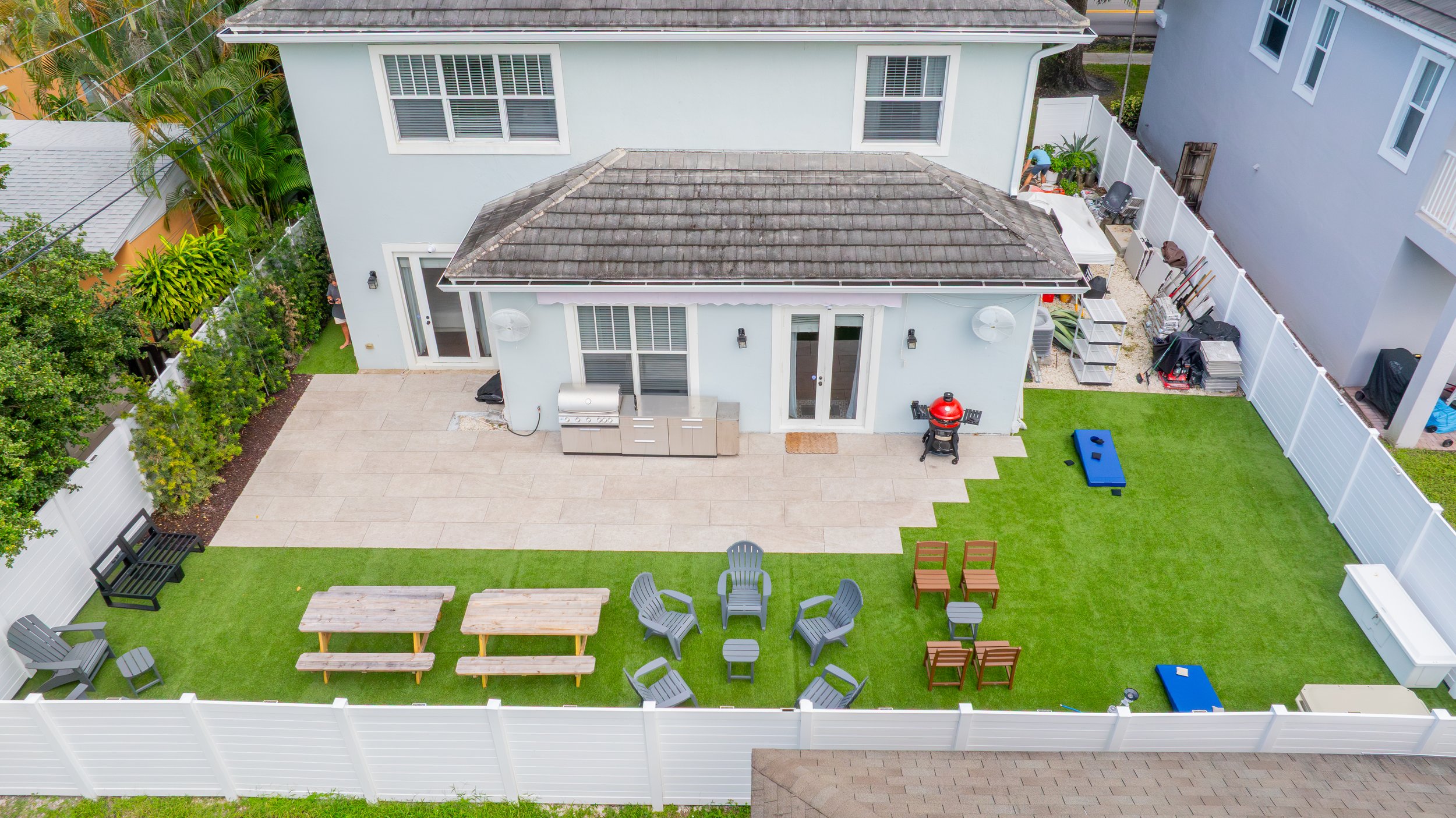 A backyard with a light blue house, a paved patio, a grassy area, various outdoor furniture, and a person working near a white fence.