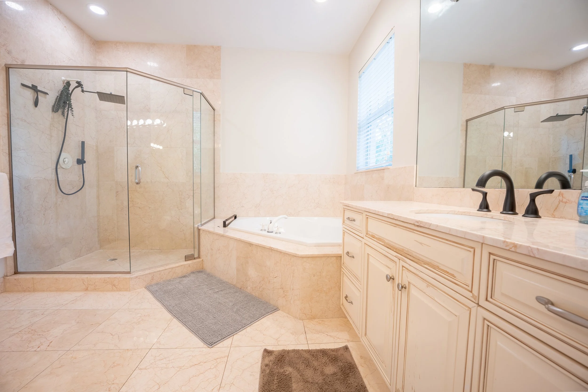 Large bathroom with a shower, bathtub, vanity with two sinks, mirror, window with blinds, and beige marble walls and floor.