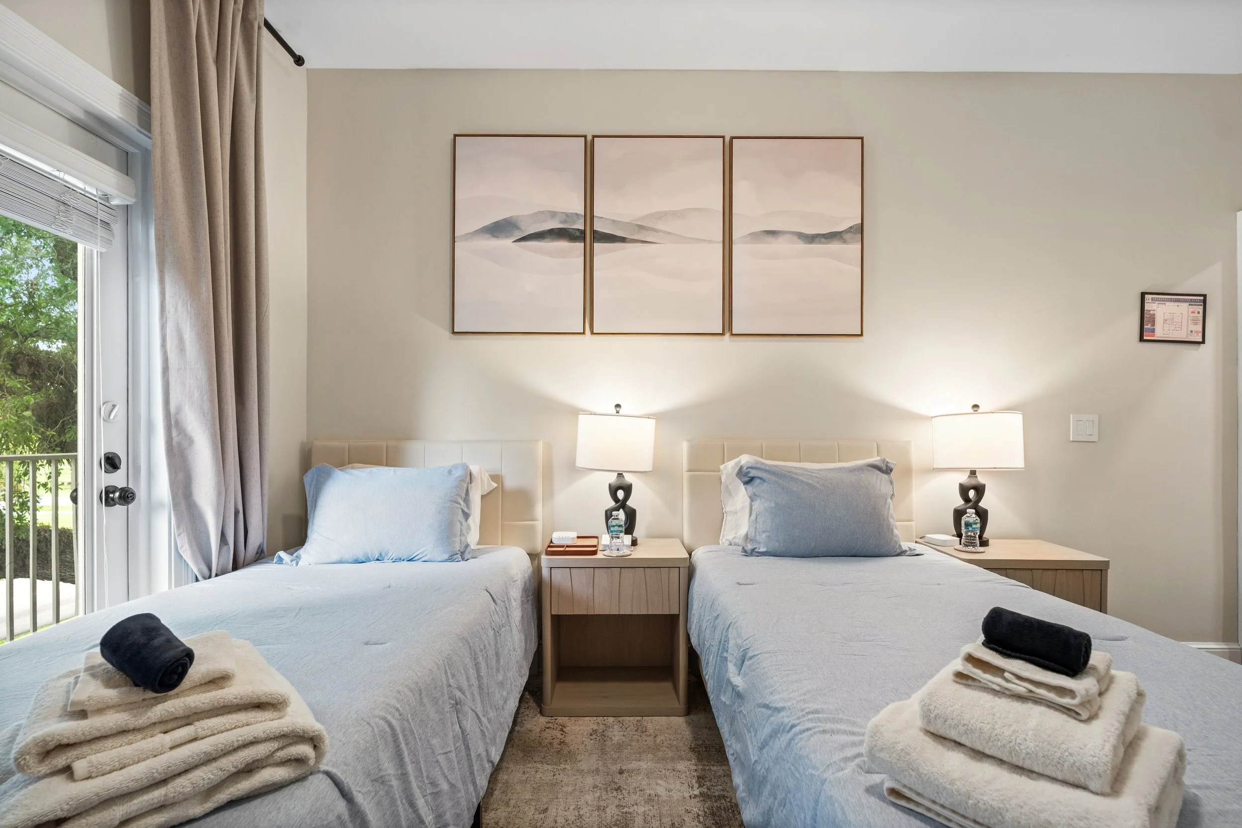 A hotel room with two beds, each with pillows and a bedside table with a lamp and water bottle, wall art above, and a window with beige curtains.