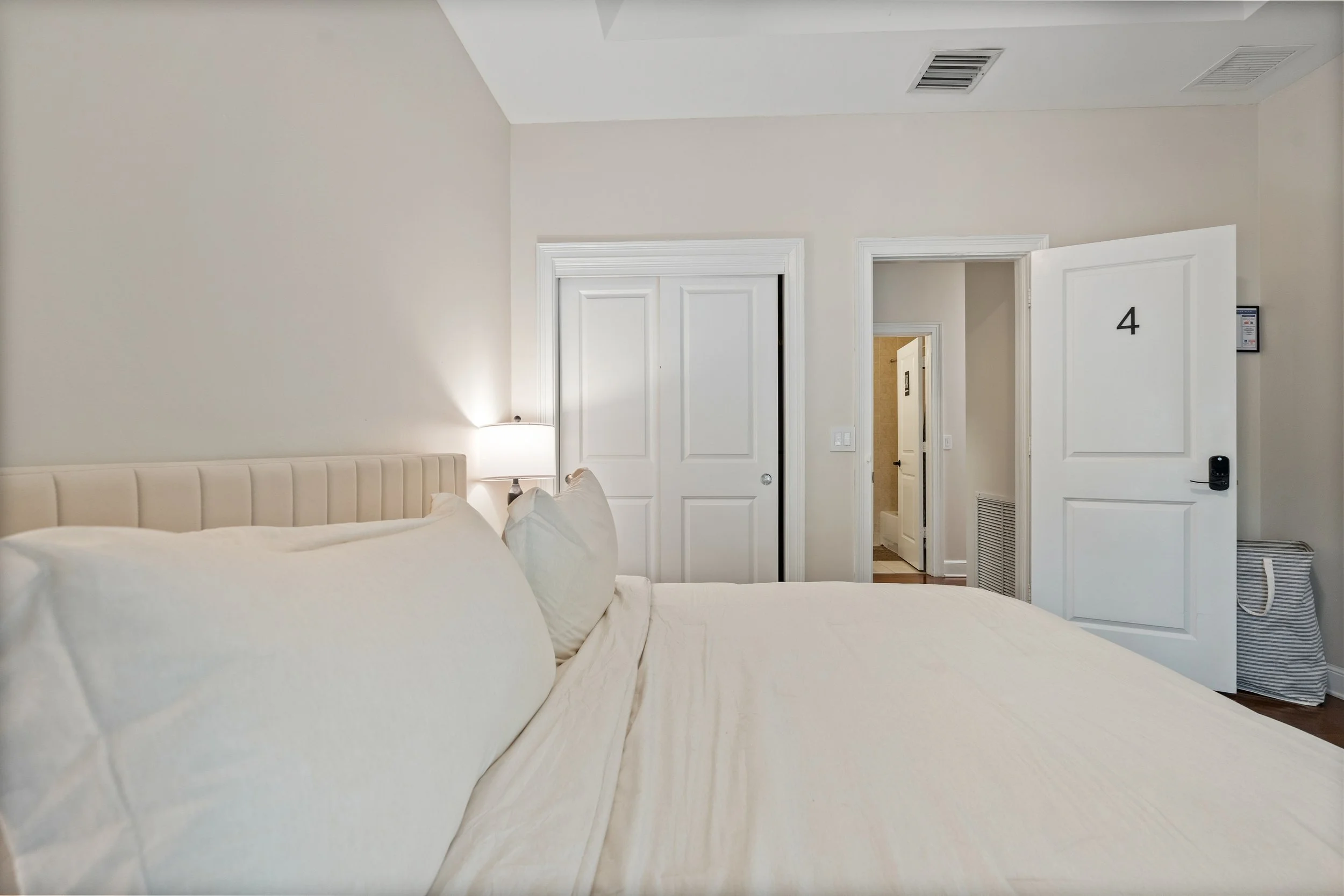 A hotel room with a bed, a lamp, two closed closet doors, an open door revealing a bathroom, and a door with the number 4. There are vents on the ceiling and a small wall-mounted device near the door.